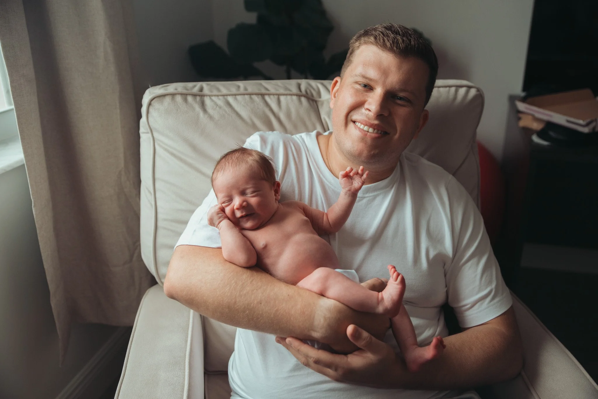 A man holding a newborn baby while sitting on a bed in a room with natural light, smiling at the camera.