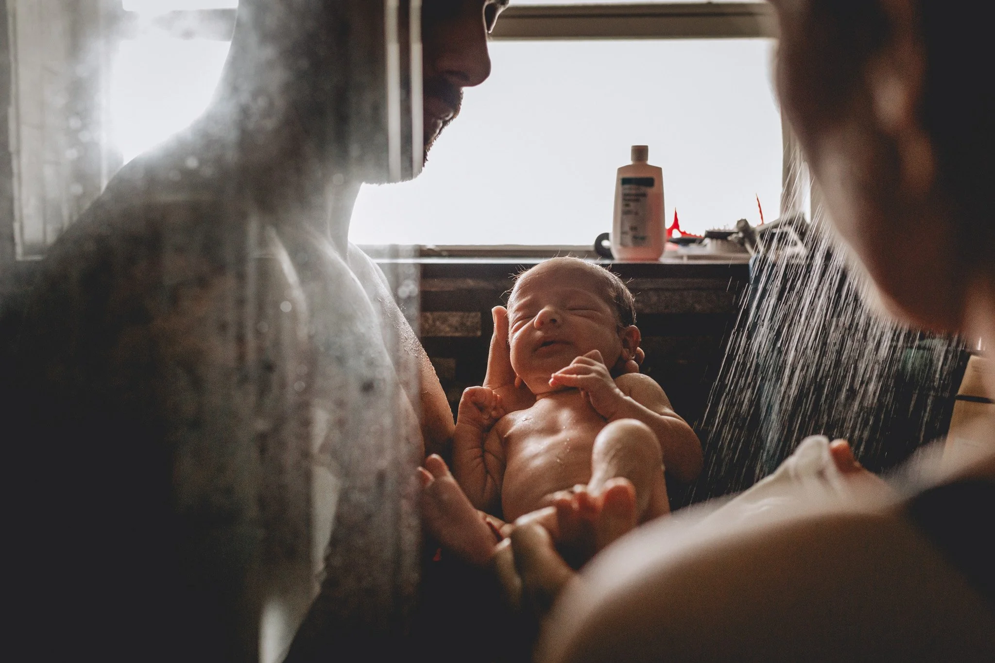 A man holding a newborn baby during a bath in a bathroom, with another person assisting. Sunlight comes through a window in the background.