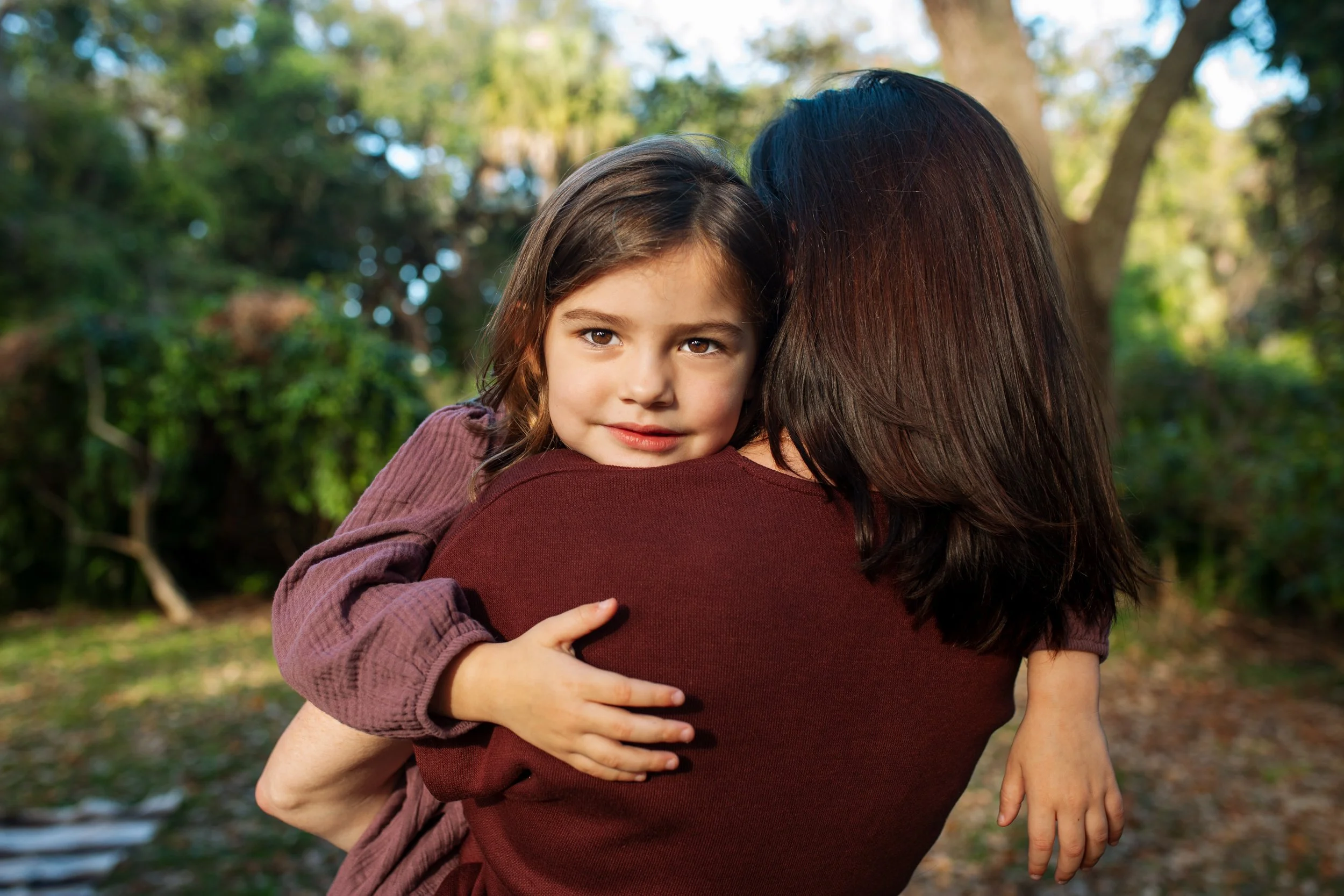 A woman holding a young girl in an outdoor park setting during daytime. The girl is looking directly at the camera with a gentle smile, while the woman is turned away, showing only her dark hair and a maroon shirt. The background consists of trees and natural greenery.