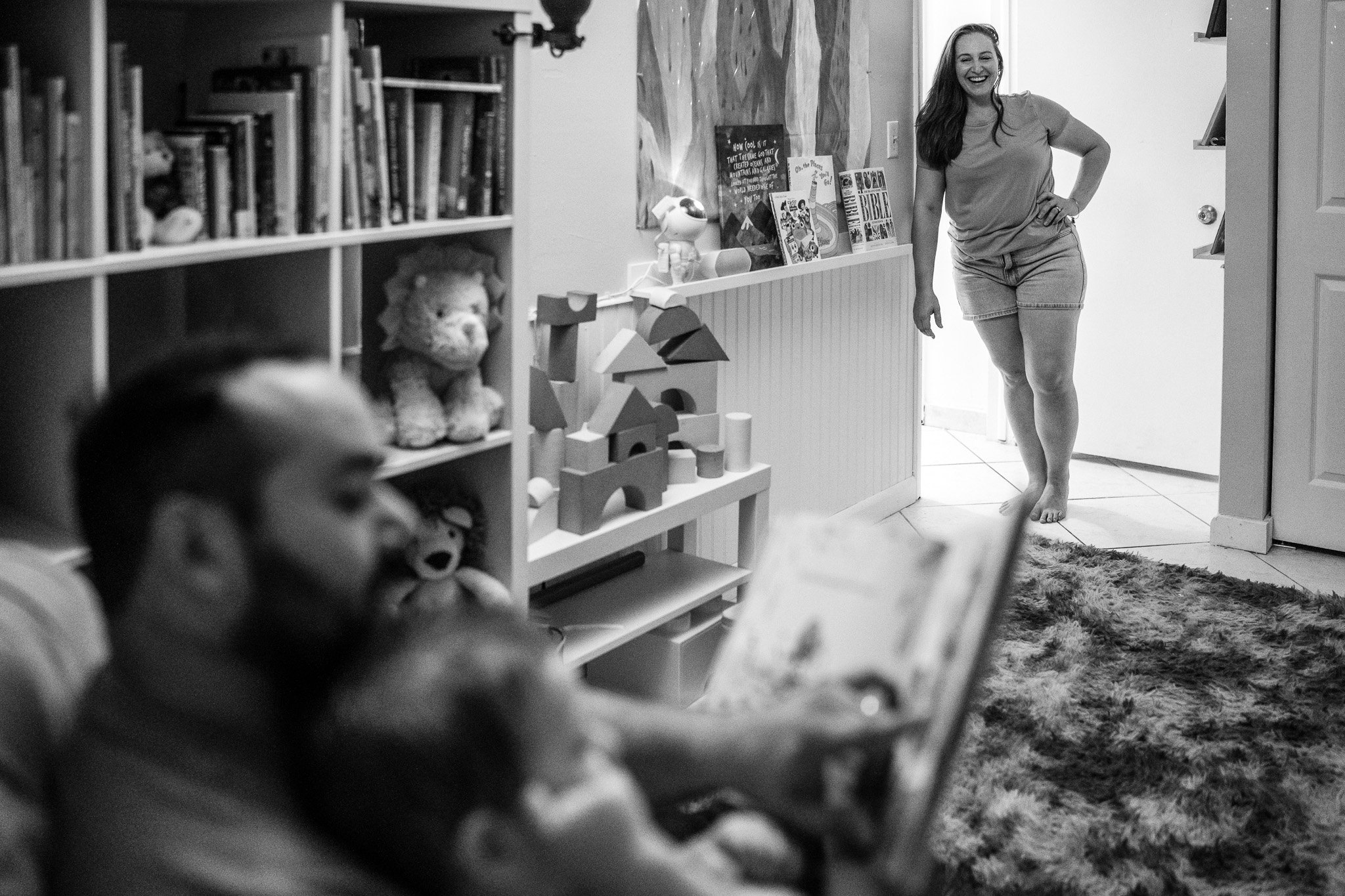 A woman smiling and standing in a doorway, with children blurred in the foreground and a bookshelf filled with toys and books behind her.