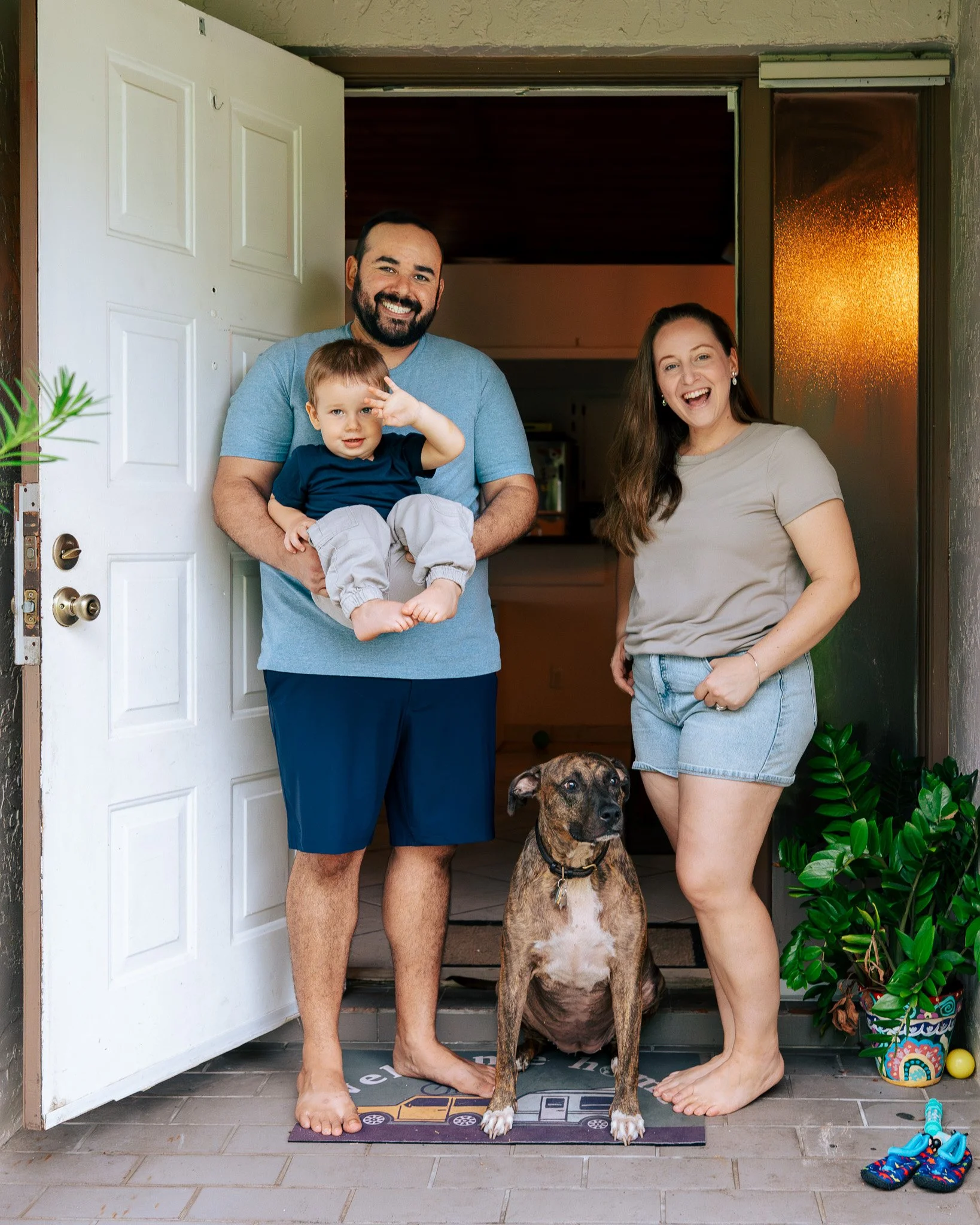 A man, woman, and young boy standing on a porch with a brindle dog, colorful shoes, and potted plant, at the entrance of a home during sunset.