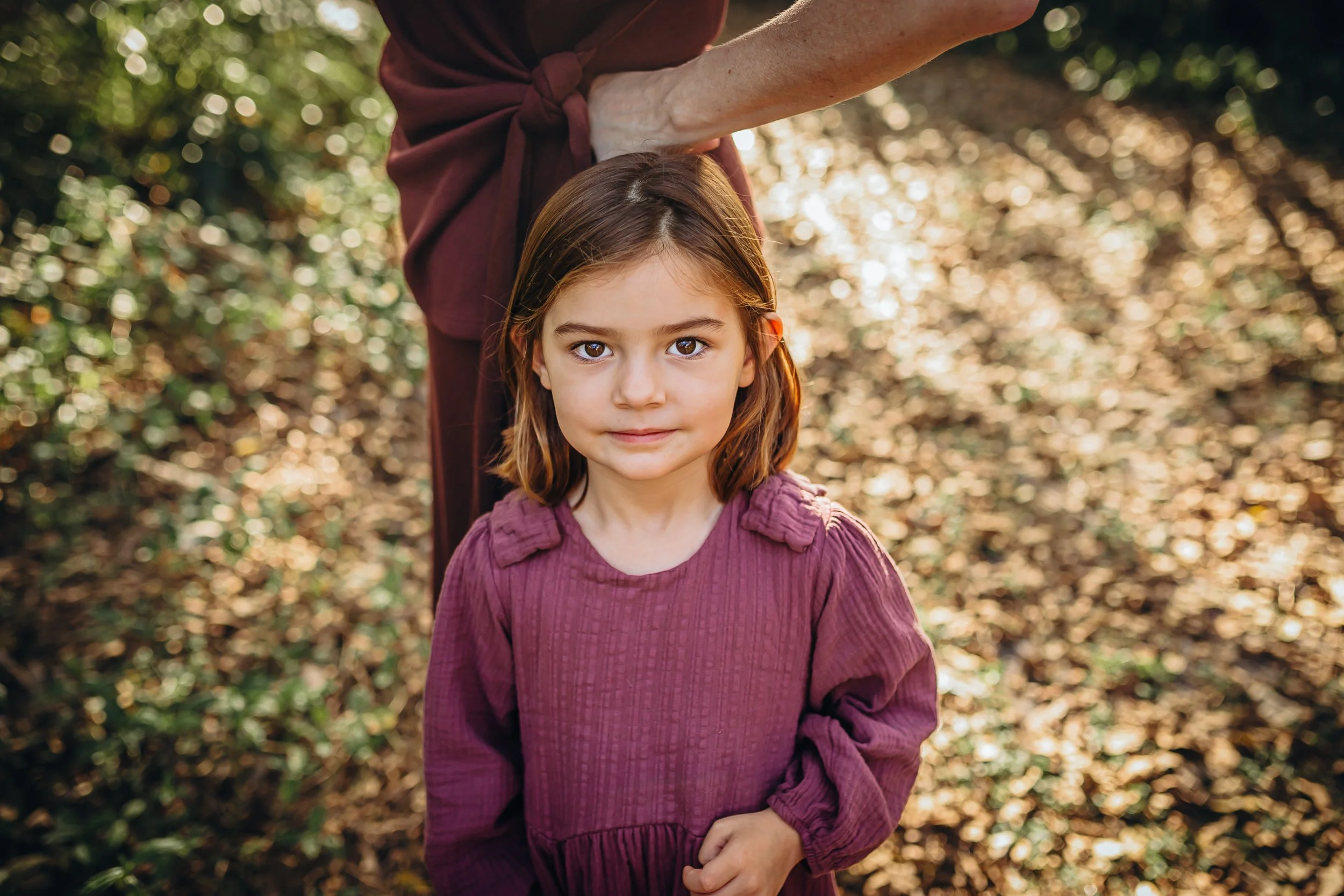 A young girl with brown hair and light skin standing in a wooded area with autumn leaves, wearing a long-sleeved mauve top. An adult, partly visible, stands behind her with one arm extended over her head, wearing a dark red top with rolled-up sleeves