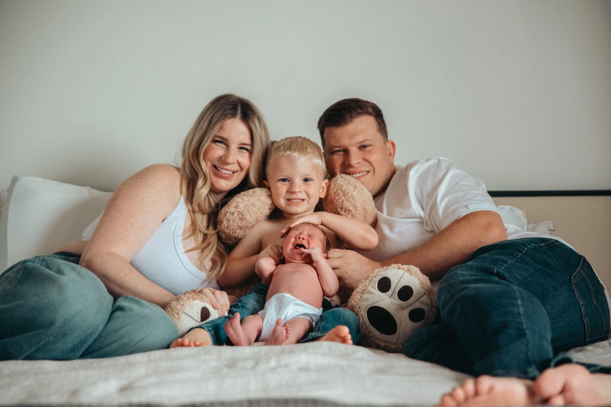 A happy family of five, including a mother, father, and three young children, sitting together on a bed and smiling at the camera.