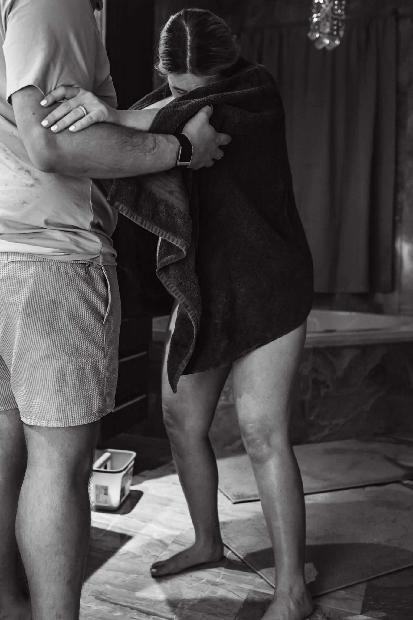 Two people, one with a tattooed arm and wearing a smartwatch, embrace a person wrapped in a dark towel or blanket. The scene appears to be indoors, with wooden paneling and some hanging decorative items in the background. The individuals are standing