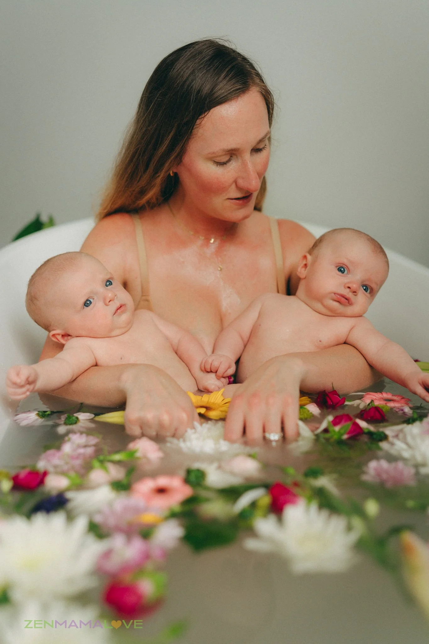 A woman with long brown hair holding two babies in a bath filled with colorful flowers and rose petals.