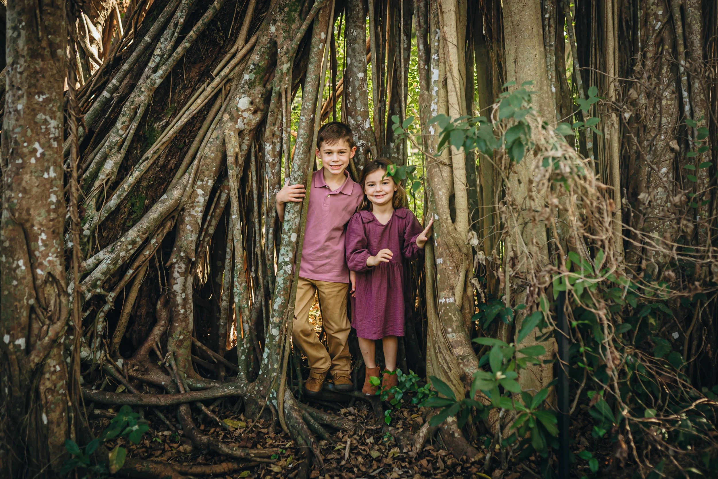 Two children, a boy and a girl, stand inside a large, natural tree structure made of intertwined branches and roots, with the boy wearing a pink shirt and khaki pants and the girl wearing a purple dress, both smiling at the camera.