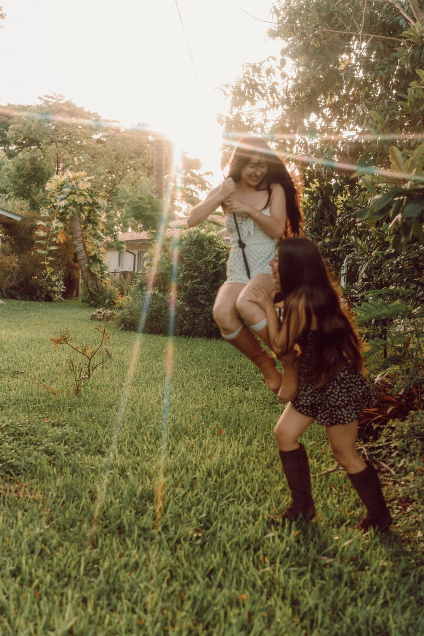 Two young women playing and laughing outdoors in a backyard with green grass, bushes, and trees, during a sunny late afternoon or early evening.
