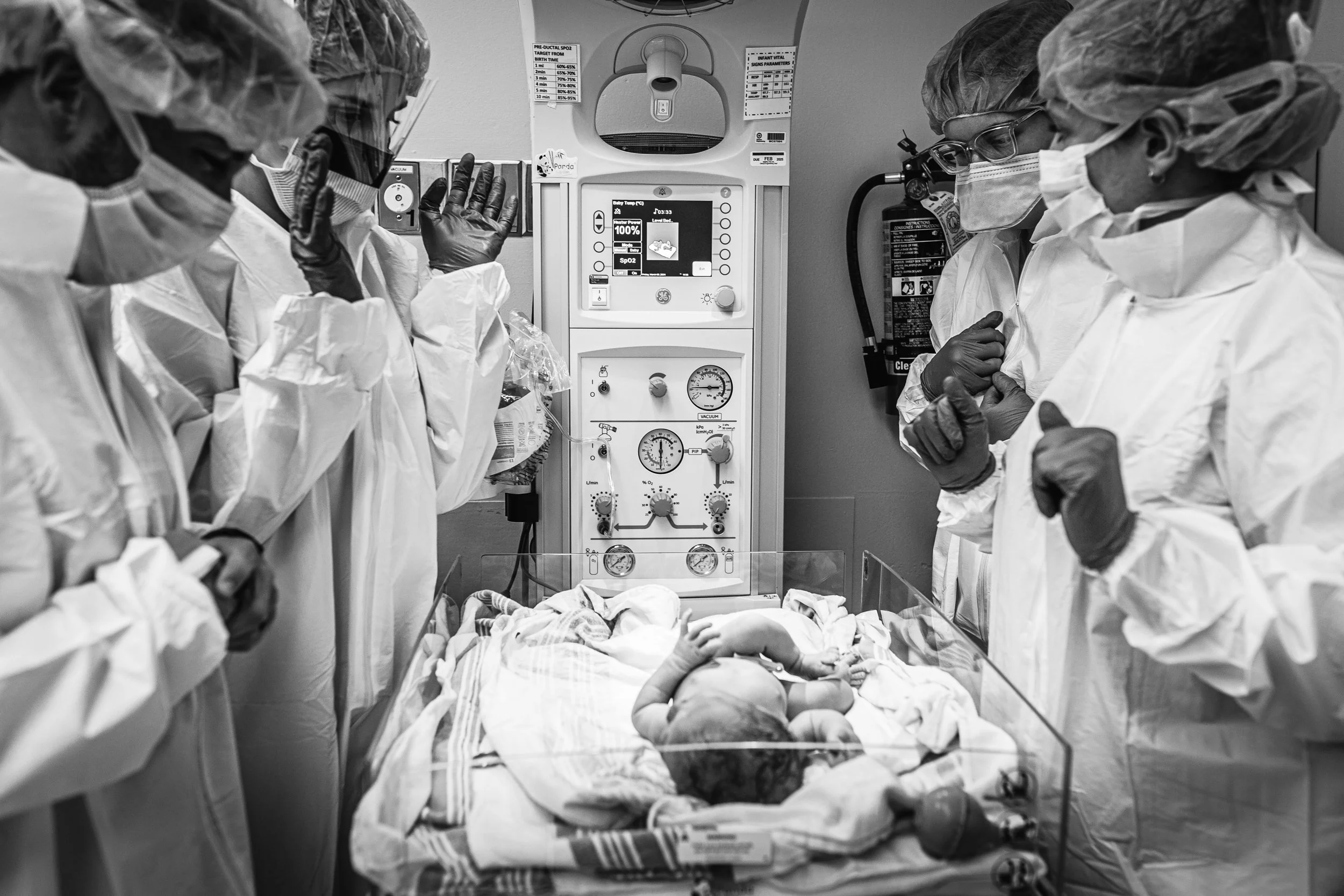 Medical professionals in protective gear surrounding a newborn in an incubator in a hospital delivery room.