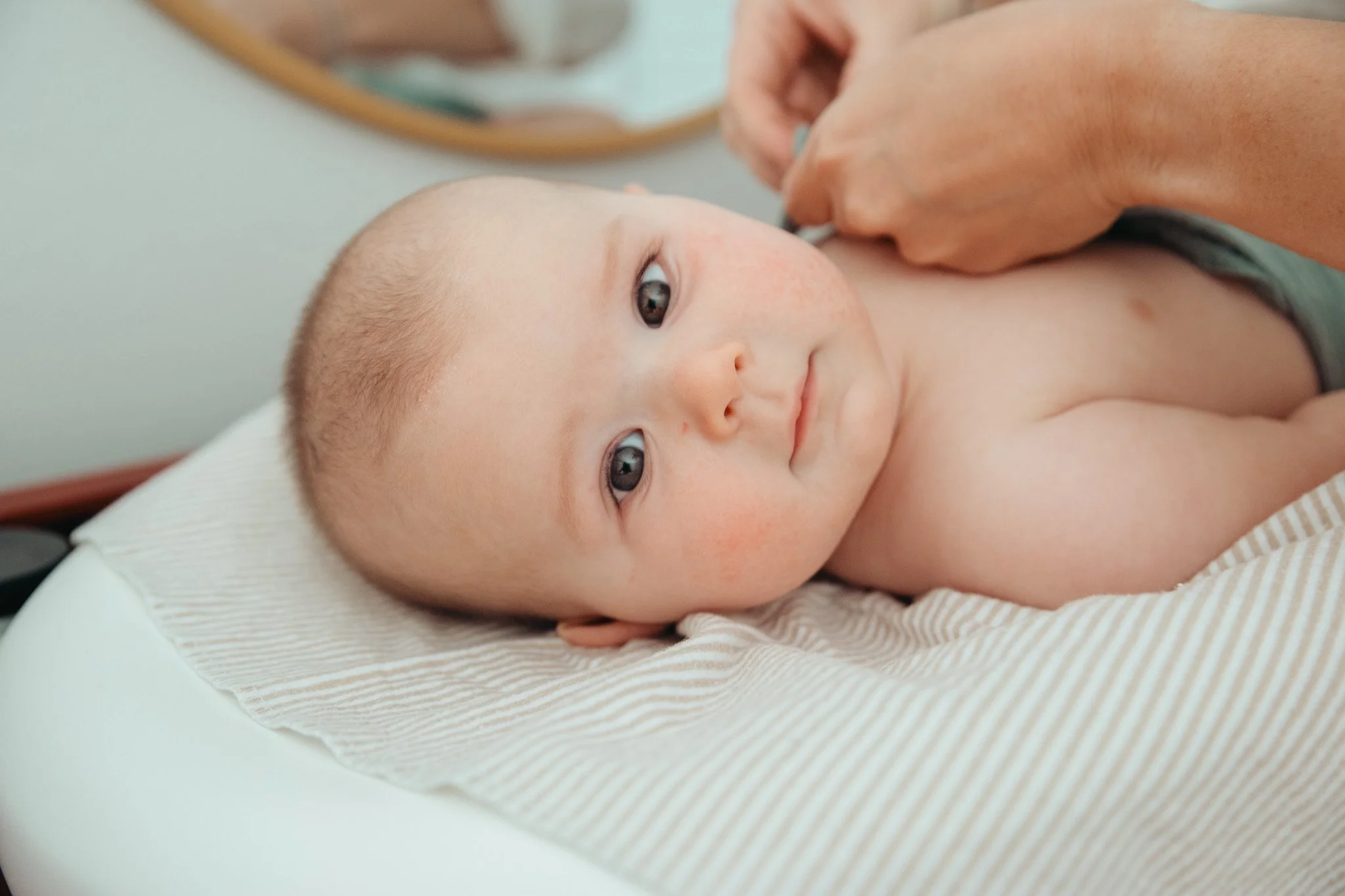 A young child lying on a bed with a white pillow, looking at the camera, while an adult is adjusting something on the child's face.