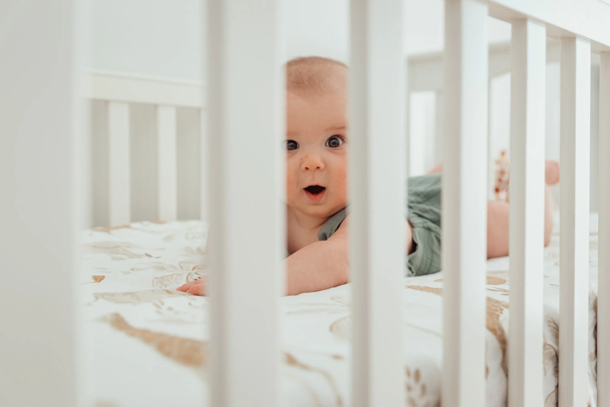 A baby lying on a bed behind white crib rails, looking through the bars with an expression of surprise or curiosity.
