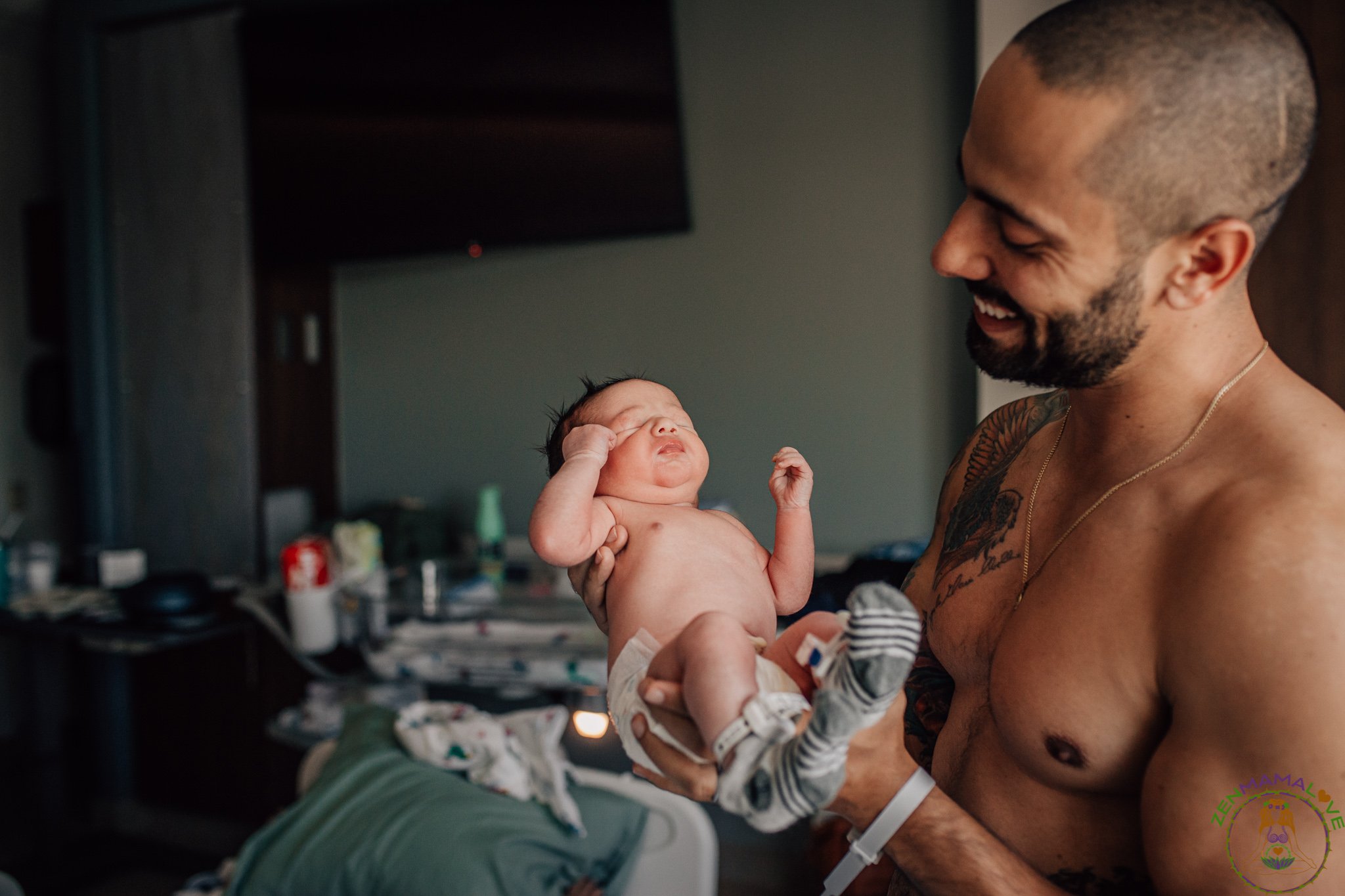 A shirtless man with tattoos holding a newborn baby in a room with a cluttered background, smiling and looking at the baby.