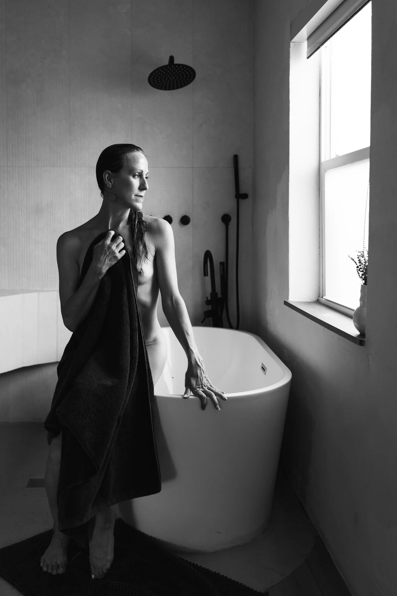 A woman with wet hair standing next to a bathtub in a bathroom, holding a towel over her shoulder, looking out the window.
