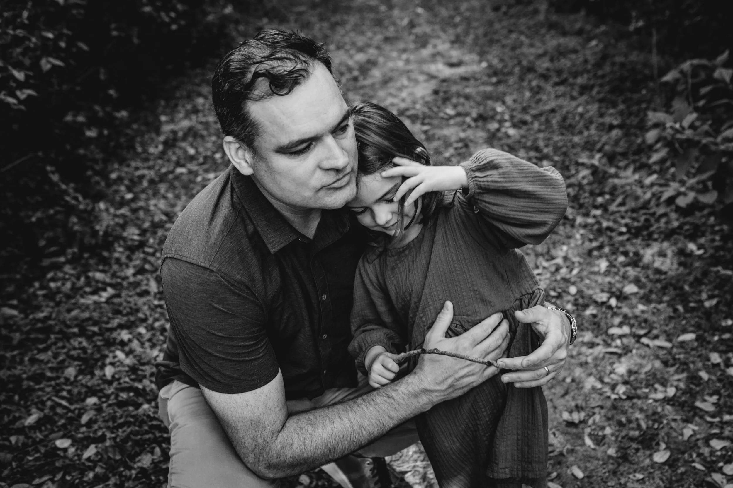 A man and a young girl are outdoors, embracing each other in a hug. The man has dark, wavy hair and is wearing a short-sleeved shirt. The girl has long hair and is wearing a long-sleeved top. The background consists of fallen leaves and forest ground