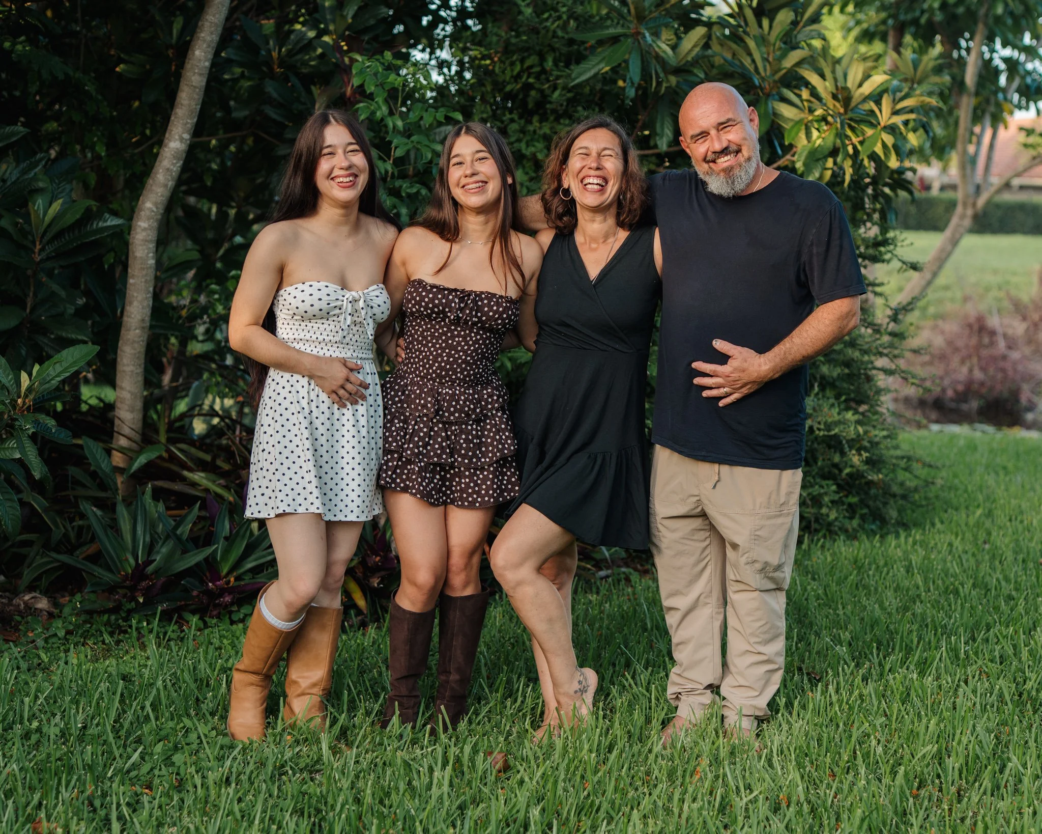 Four people standing outdoors in a garden, smiling and laughing, with lush green plants and trees in the background.