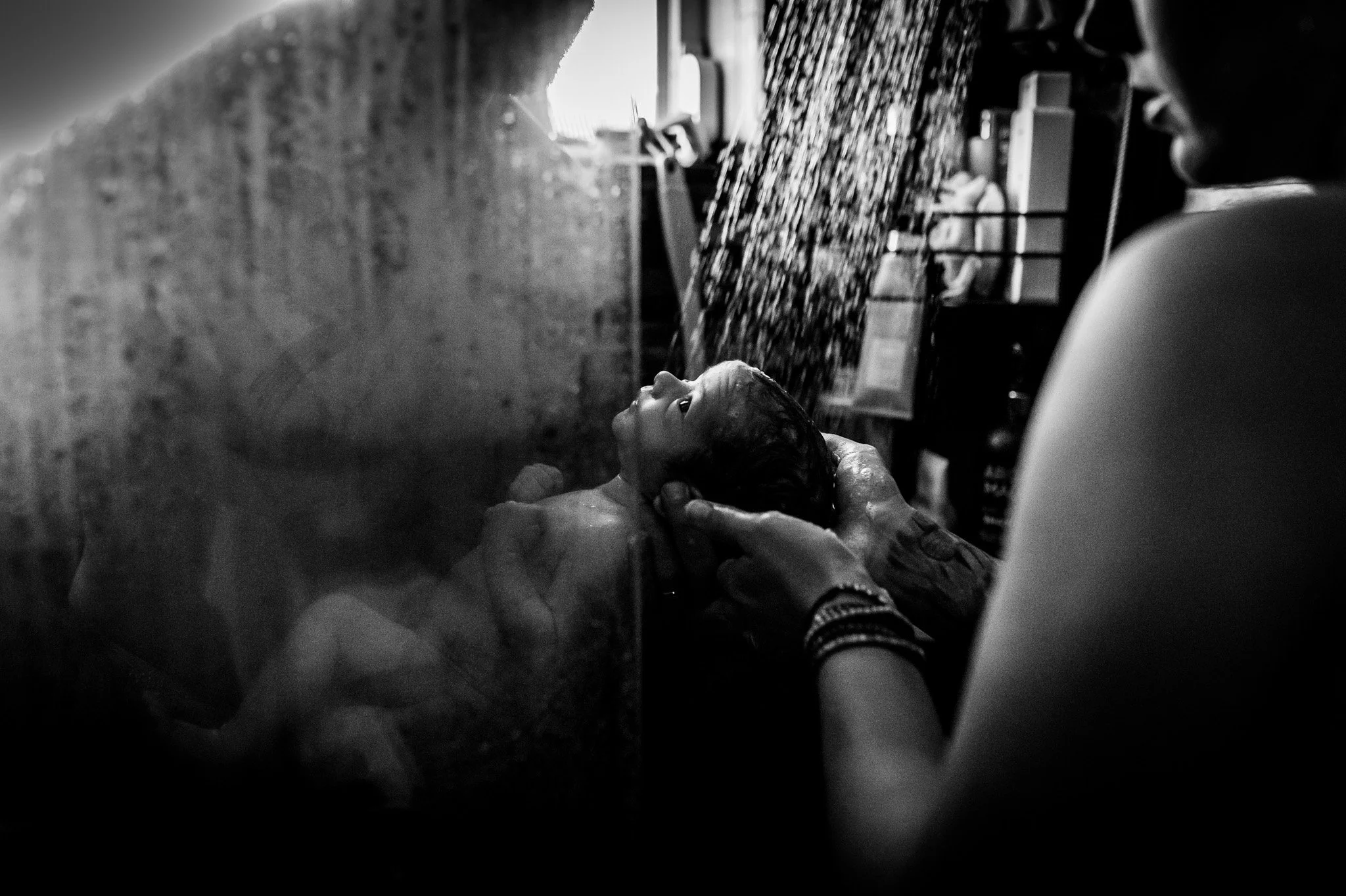 A person giving a bath to a young girl in a bathroom, with water pouring over her head, viewed through a glass shower door.