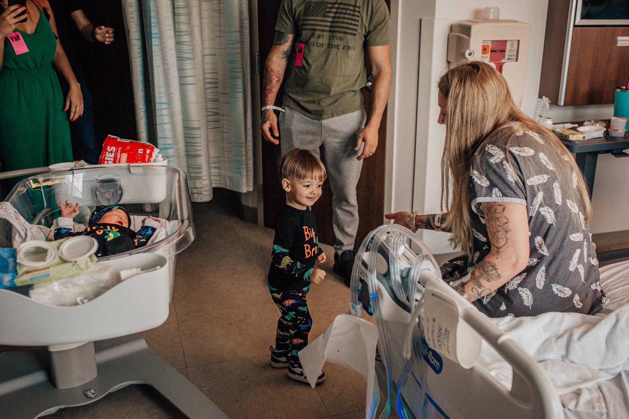 A hospital room with a newborn baby in an incubator, a woman with tattoos leaning over the incubator, a young boy smiling and standing nearby, and several adults standing around. Medical equipment and supplies are visible, with a curtain and a water 