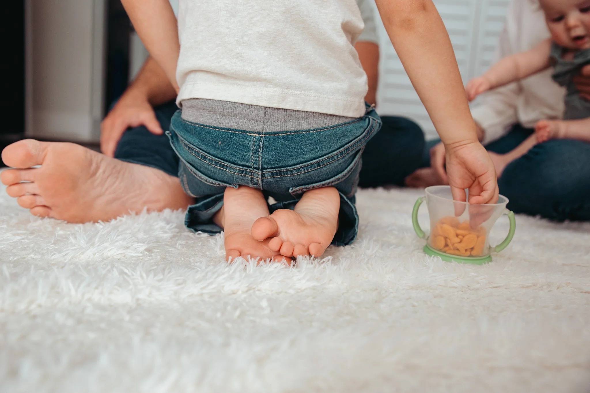 Children sitting on a fluffy white carpet, with one child in jeans and a light shirt, playing with a clear cup filled with snacks, while another child in the background is reaching towards the cup.