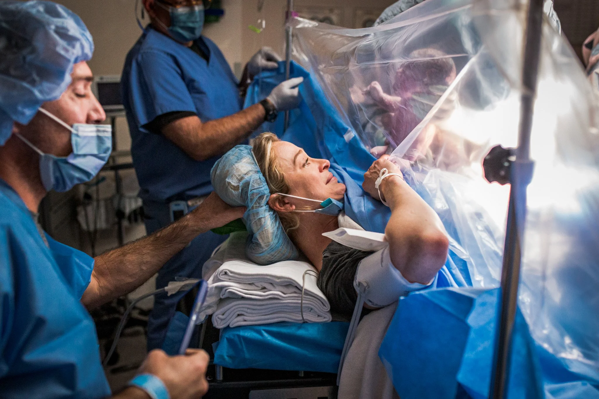 Medical team in protective gear performing a procedure on a female patient lying on a hospital bed, in an operating room or isolation setting.