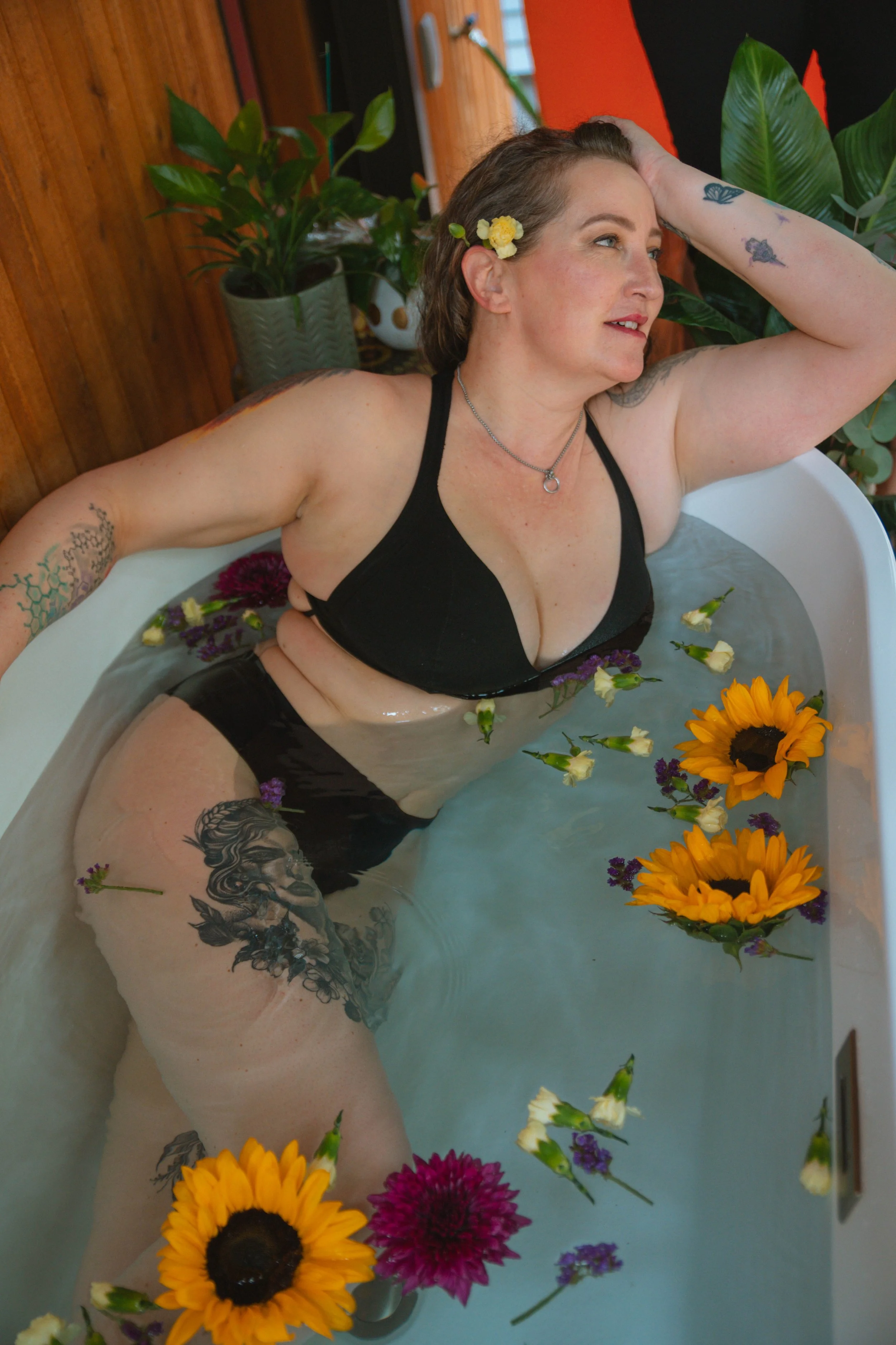 A woman relaxing in a bathtub filled with water and surrounded by colorful flowers, with her arm resting behind her head and looking content.