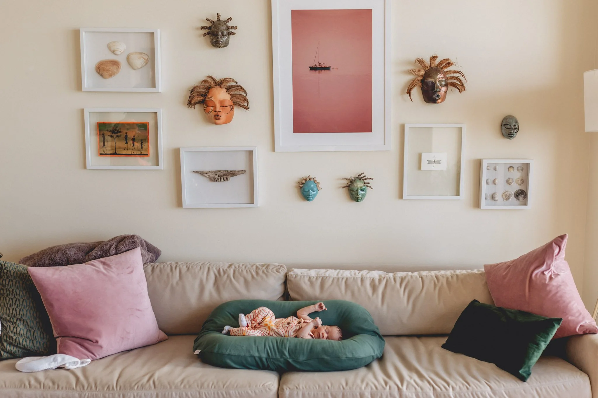 Living room with beige sofa, pink and black pillows, wall art including masks, a photograph of a boat at sea, and shells, with a baby lying on a green cushion in front of the sofa.
