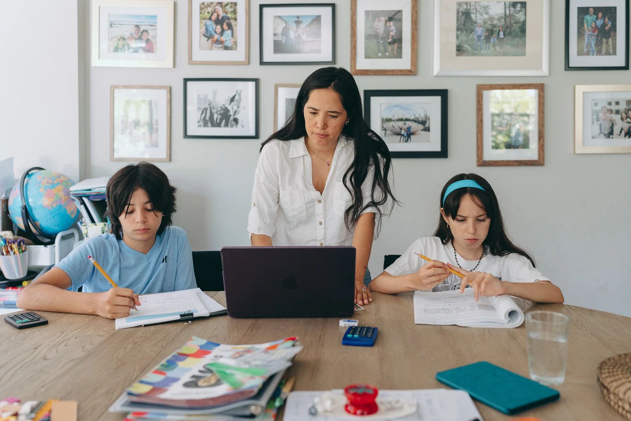 A teacher supervising two students during class at a desk covered with books, papers, and school supplies, with framed photographs hanging on the wall behind them.