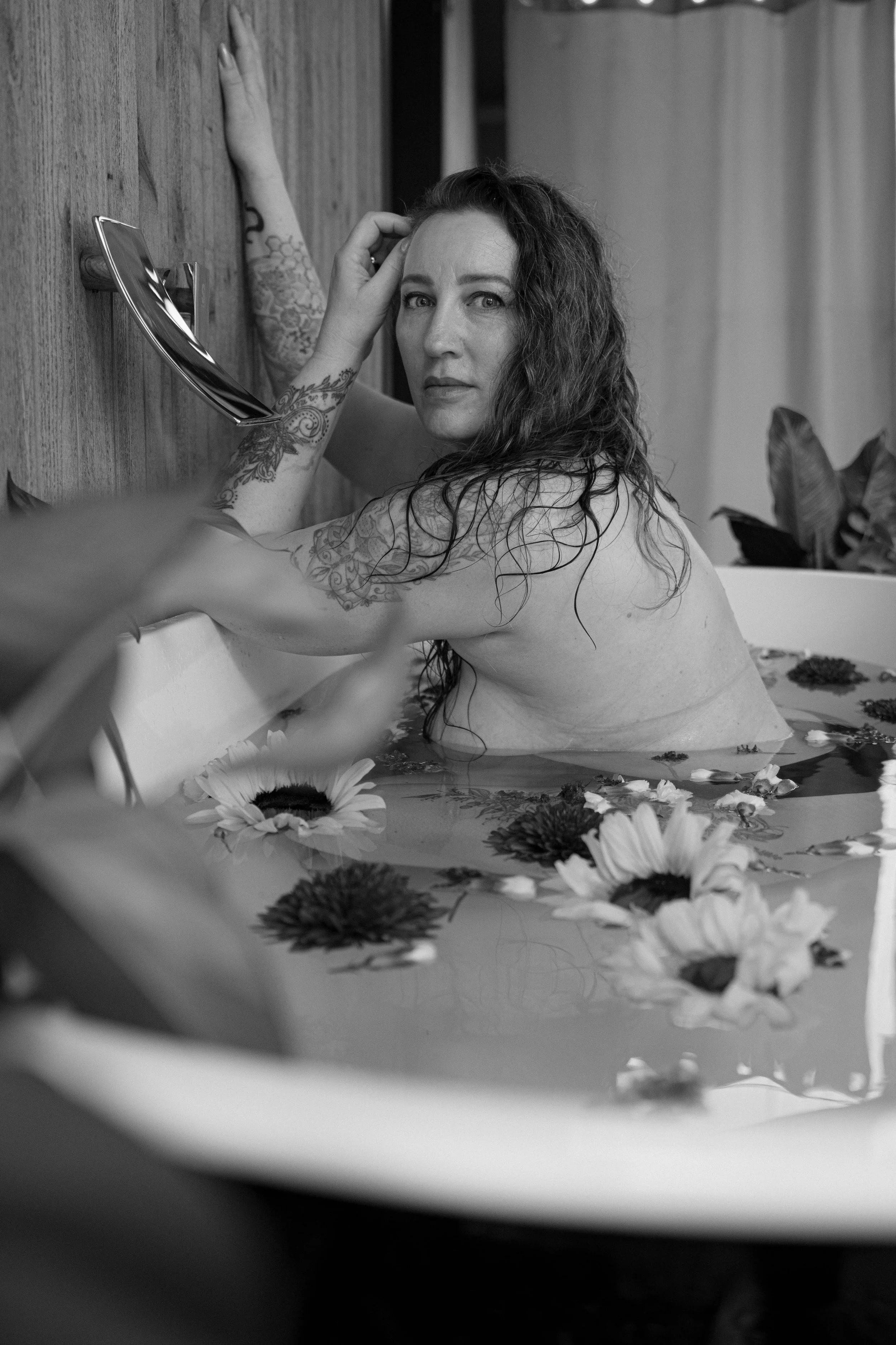A woman with long curly hair and tattoos on her arm, sitting in a bathtub filled with floating flowers, looking at the camera.