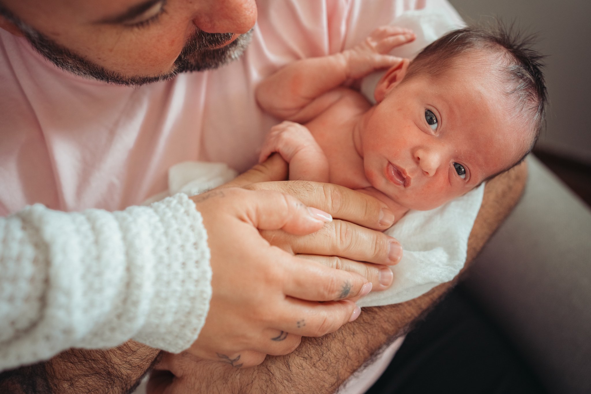 A man holding a newborn baby close to his chest, gently supporting the baby's body with his hands, while the baby looks at the camera with wide, curious eyes.