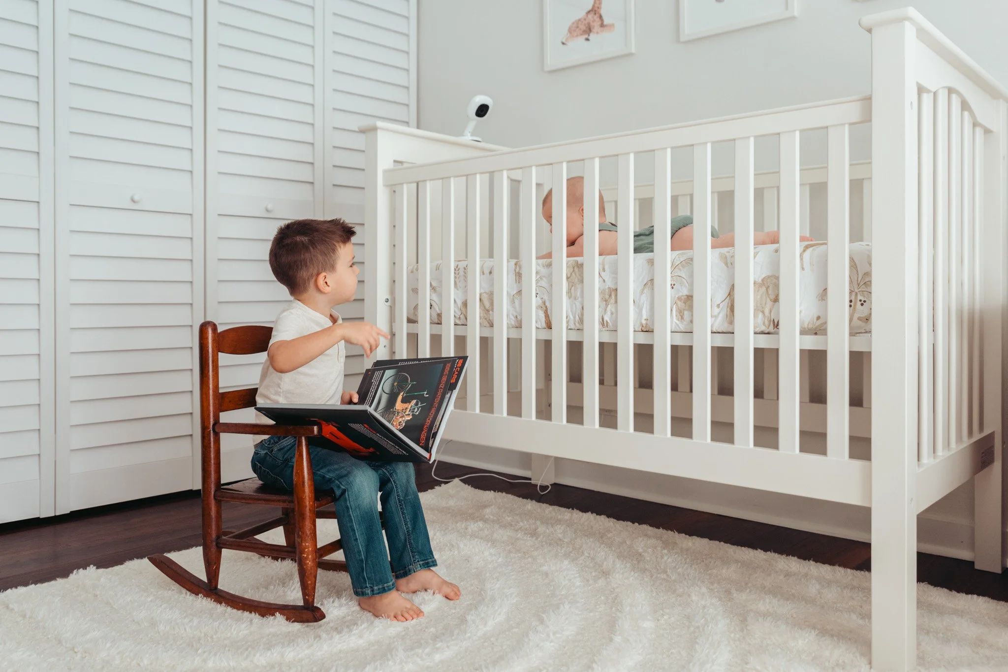 A young boy sitting on a wooden rocking chair reading a book, while watching a baby in a crib with a security camera mounted above.
