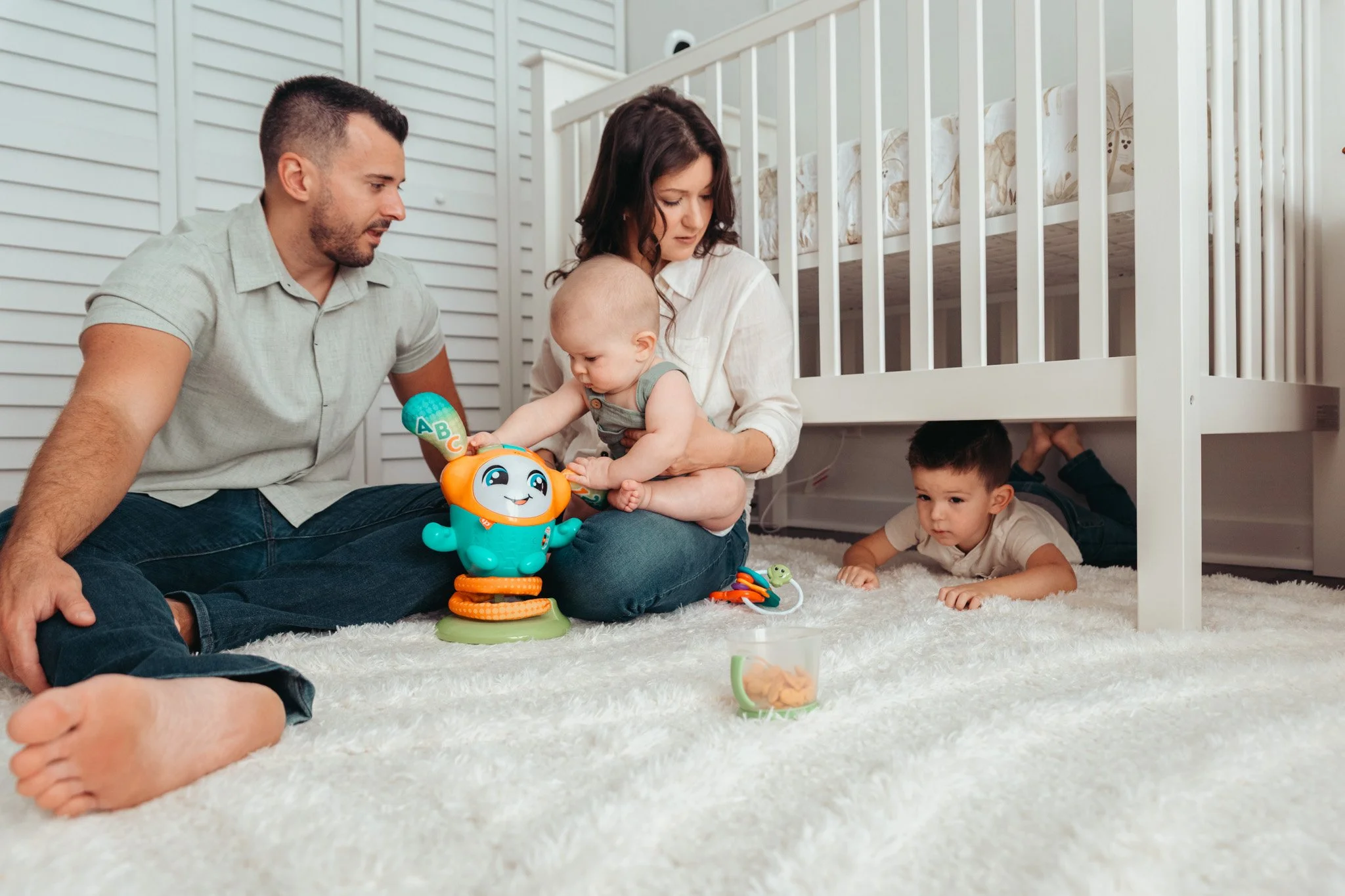 Family of four playing and relaxing on a white carpet in a nursery, with a crib in the background. The mother is holding a baby who is playing with a colorful toy, while the father watches. A young boy is lying on the floor, and a toddler is sitting 