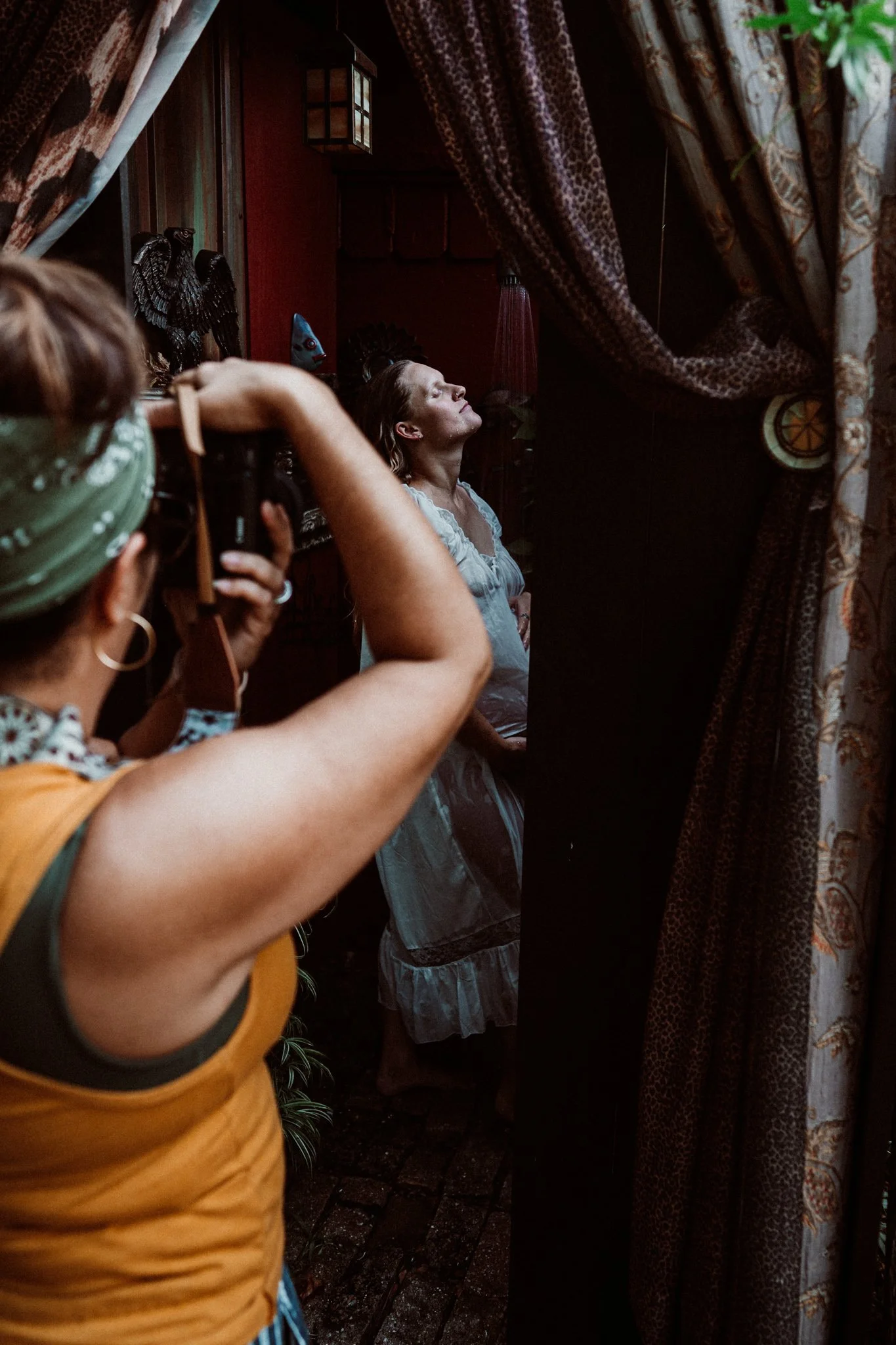 Person with striped shirt and styled hair posing in a doorway with colorful curtains, antique decor, and a woman with floral top taking a photo.