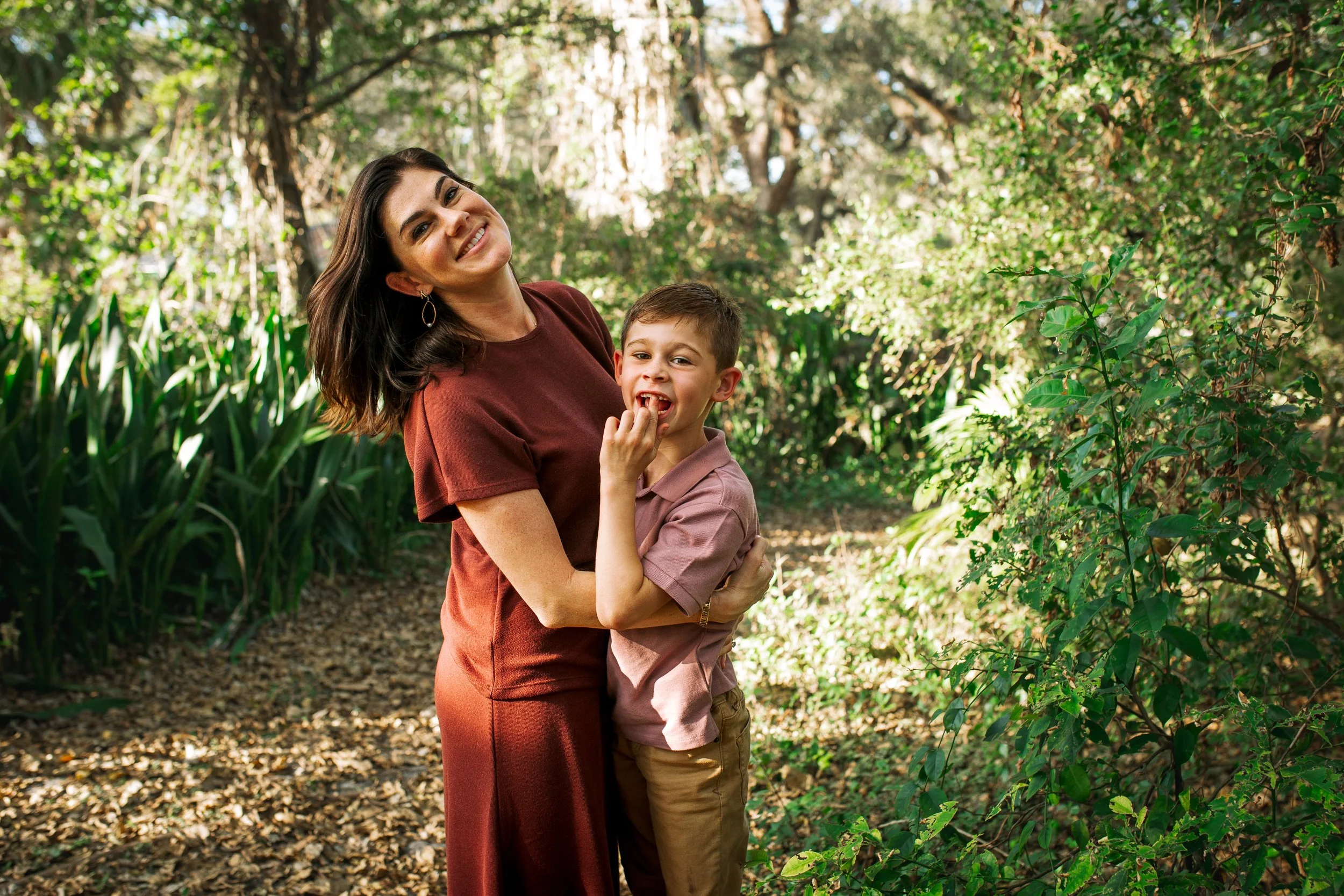 A woman and a boy smiling and hugging each other in a wooded park with sunlight filtering through the trees.