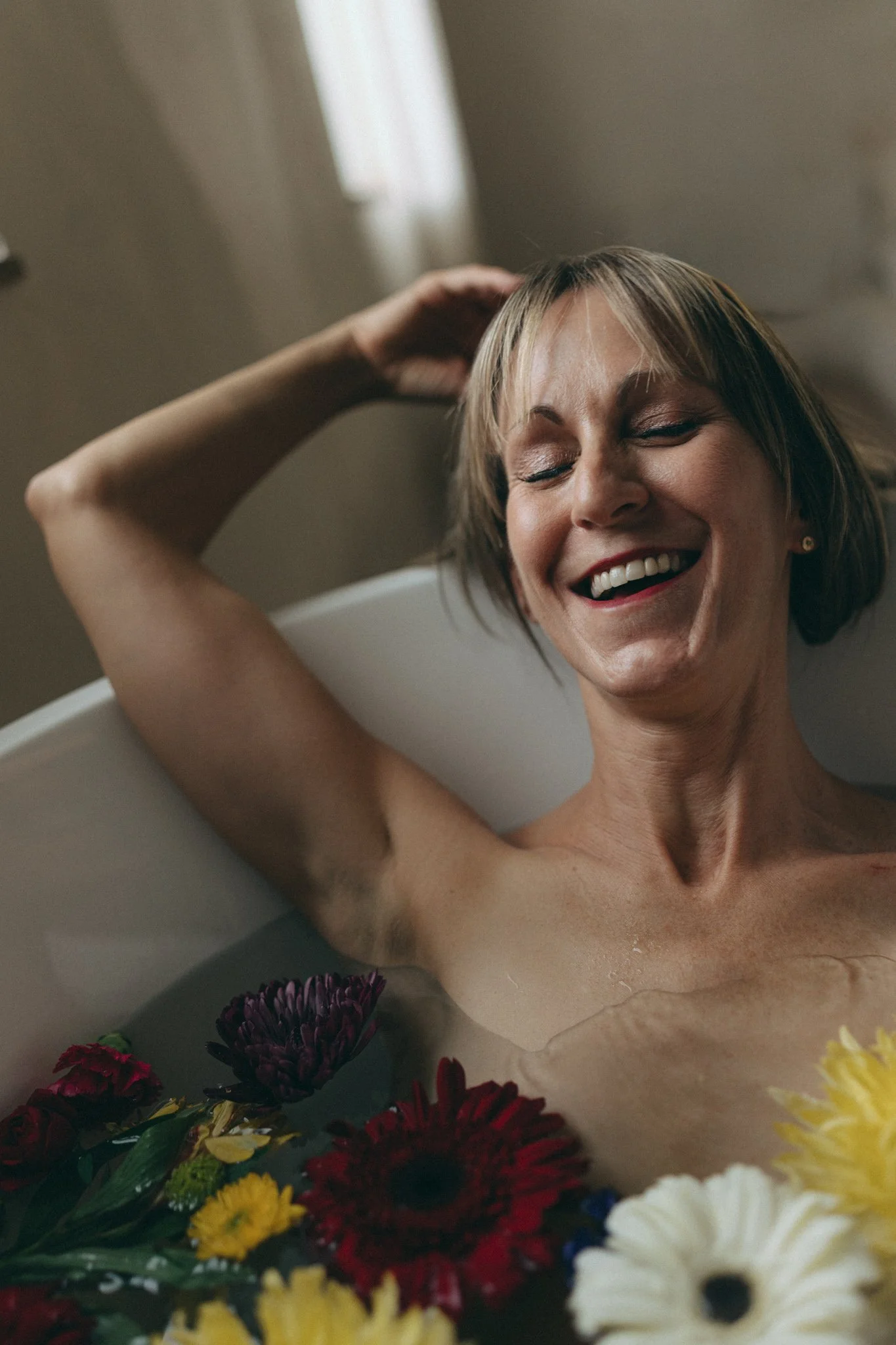 A woman with short hair, smiling with eyes closed, lying in a bathtub surrounded by colorful flowers.