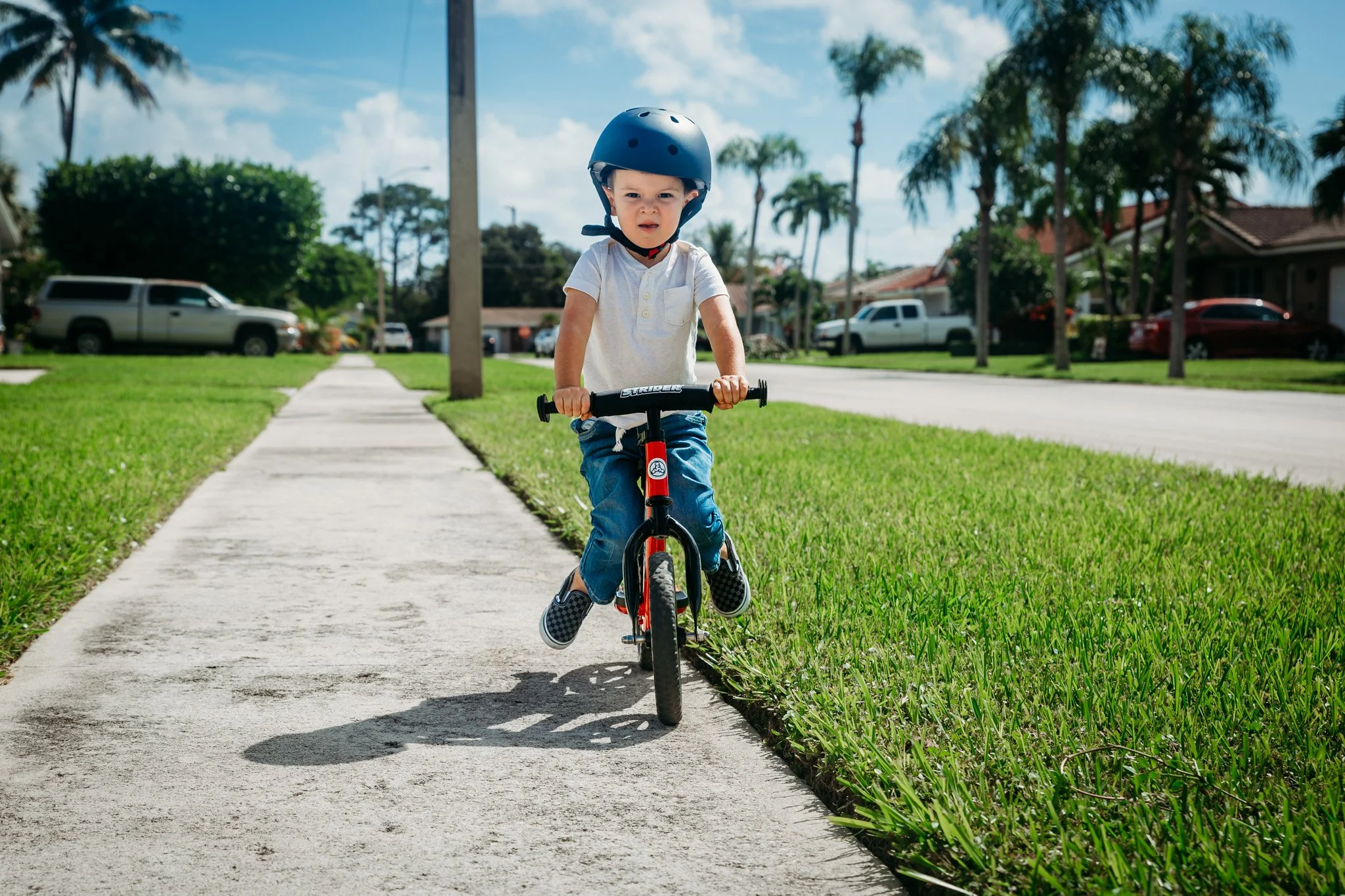 A young boy riding a bicycle on the sidewalk in a suburban neighborhood, wearing a helmet, with houses, palm trees, and parked cars in the background.