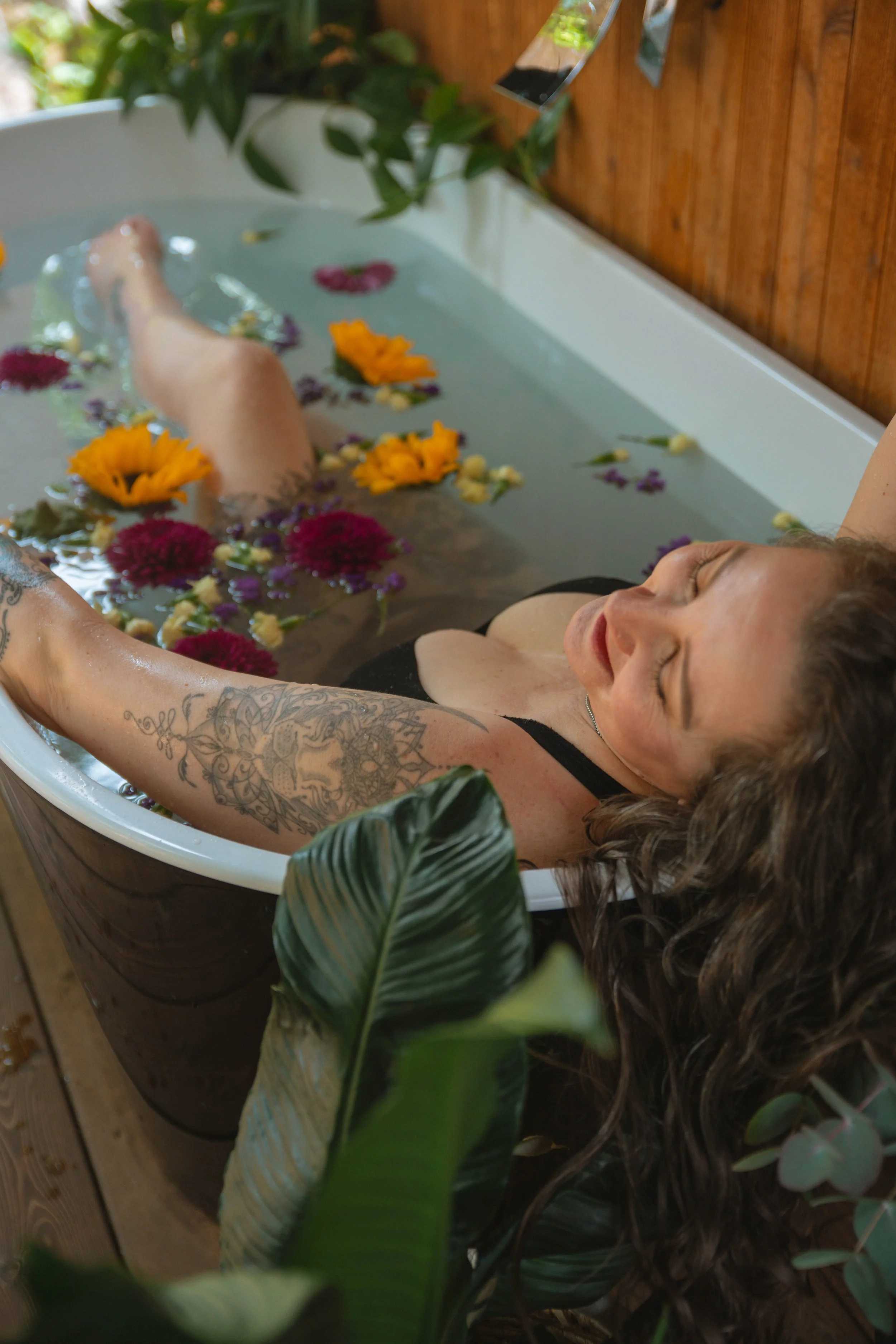 A woman with tattoos on her arms and long curly hair is relaxing in a bathtub filled with water and colorful flower petals. The bathtub is positioned on a wooden deck with greenery nearby.