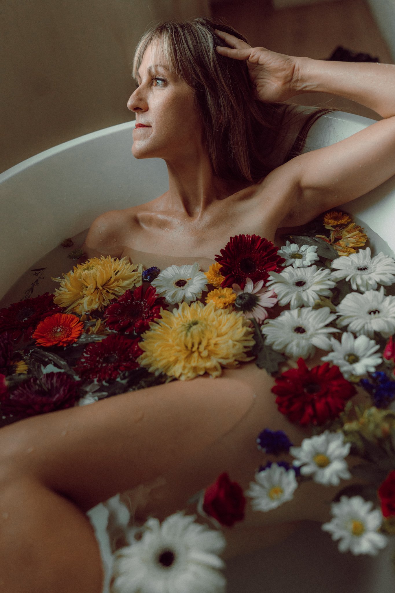 A middle-aged woman with brown hair and blue eyes relaxing in a bathtub filled with various colorful flowers, looking to the side with a peaceful expression.