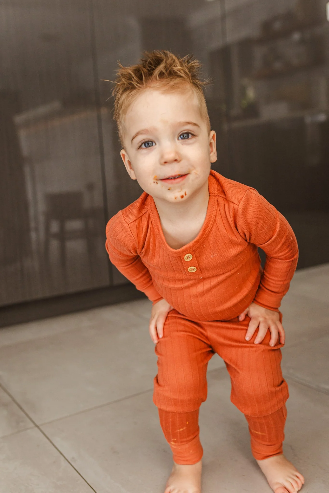 A young boy with spiky light brown hair, blue eyes, and light skin, wearing an orange outfit with buttons, standing on a tiled floor in front of a dark-colored cabinet, with some food remnants around his mouth.