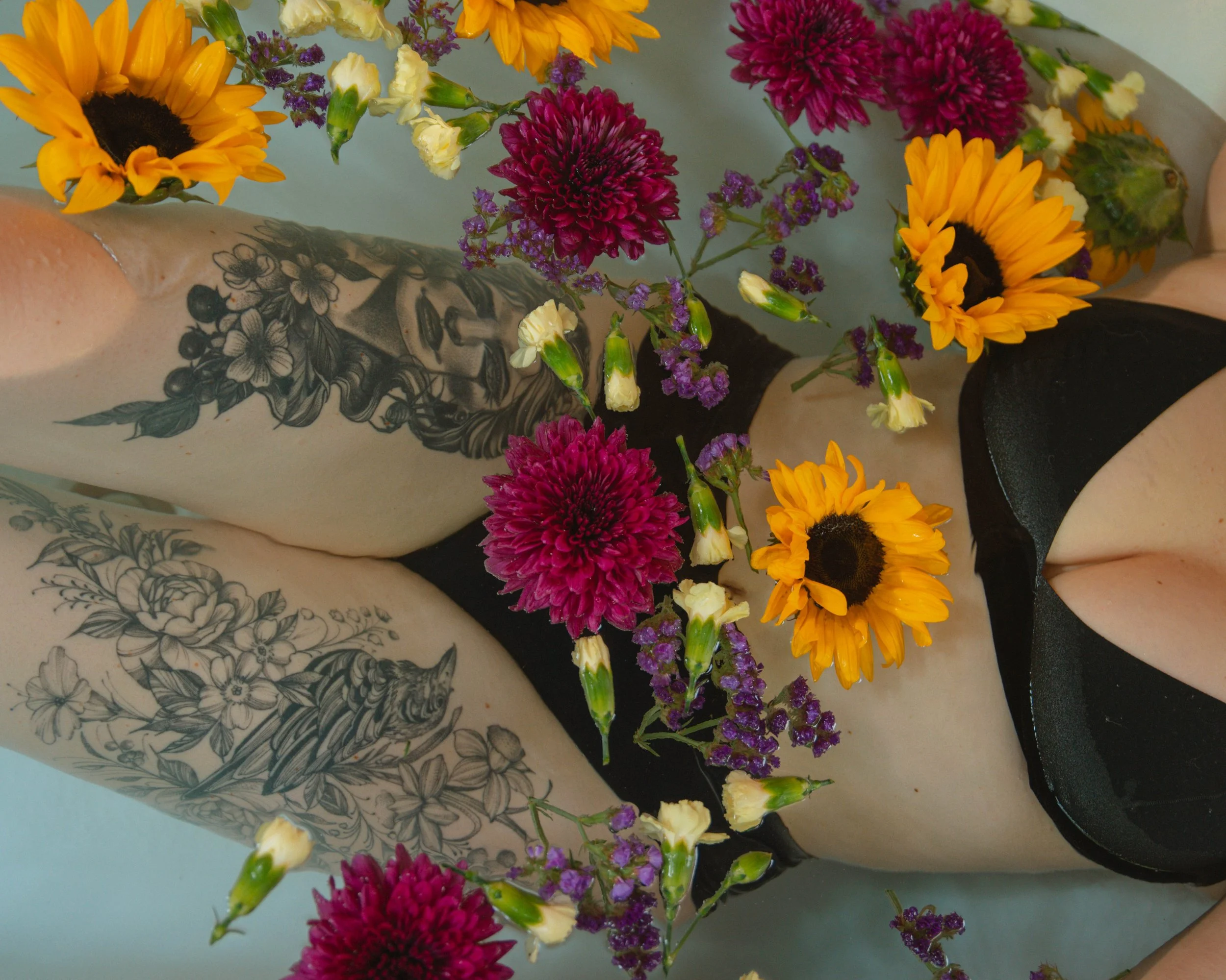 Colorful flowers including yellow sunflowers, purple asters, and small yellow and white blooms floating in a bathtub or similar container with tattoos visible on the person's arm.