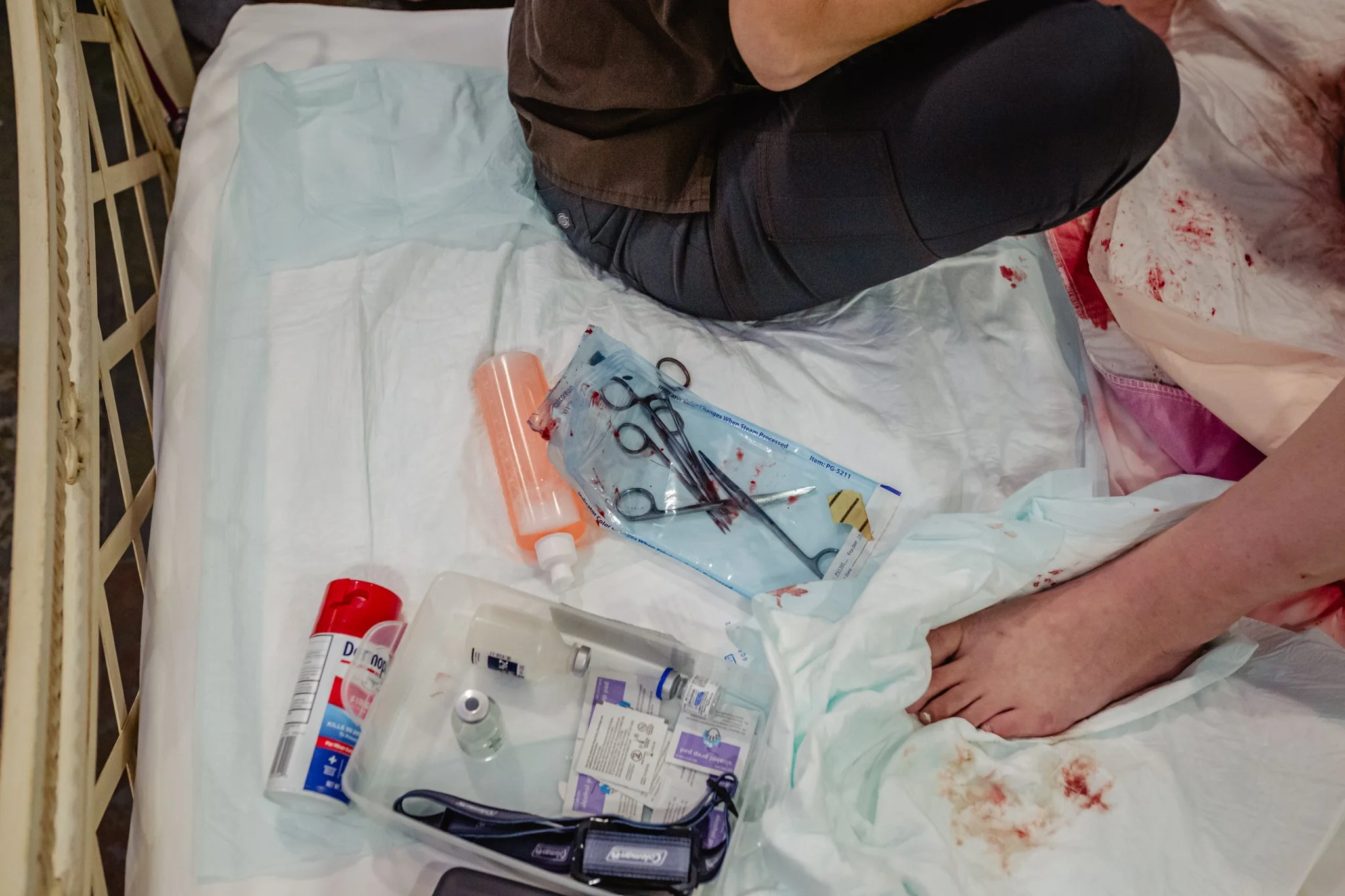 Scene of a hospital bed with medical supplies including scissors, syringe, medication bottles, and other medical equipment, and a person sitting on the edge of the bed. There are blood stains on the bed's sheet and surrounding area.