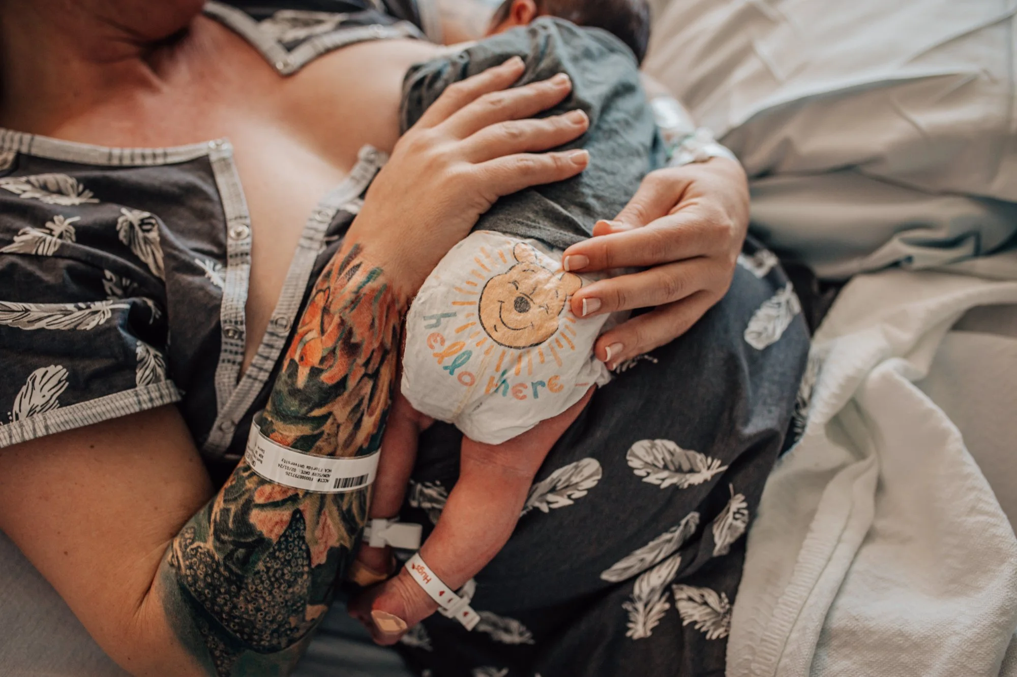 A woman holding a newborn baby in her arms, with medical wristbands on both, lying in a hospital bed. The baby wears a diaper with a cheerful Winnie the Pooh graphic and the words 'You're here', and is wrapped in a blanket. The woman's tattoos and fl