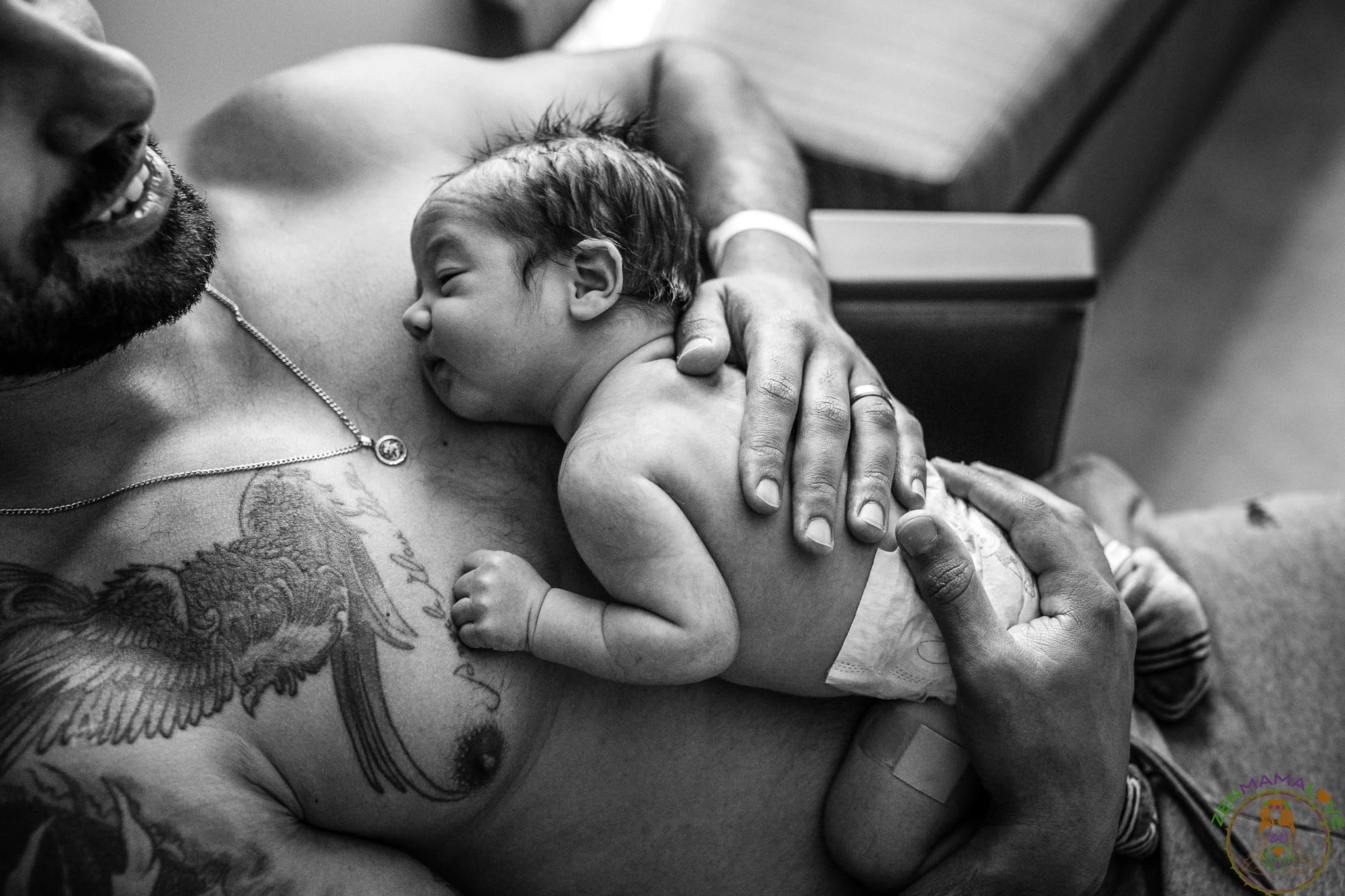 A man with tattoos on his arm and chest holding a newborn baby close to his chest in a hospital room.
