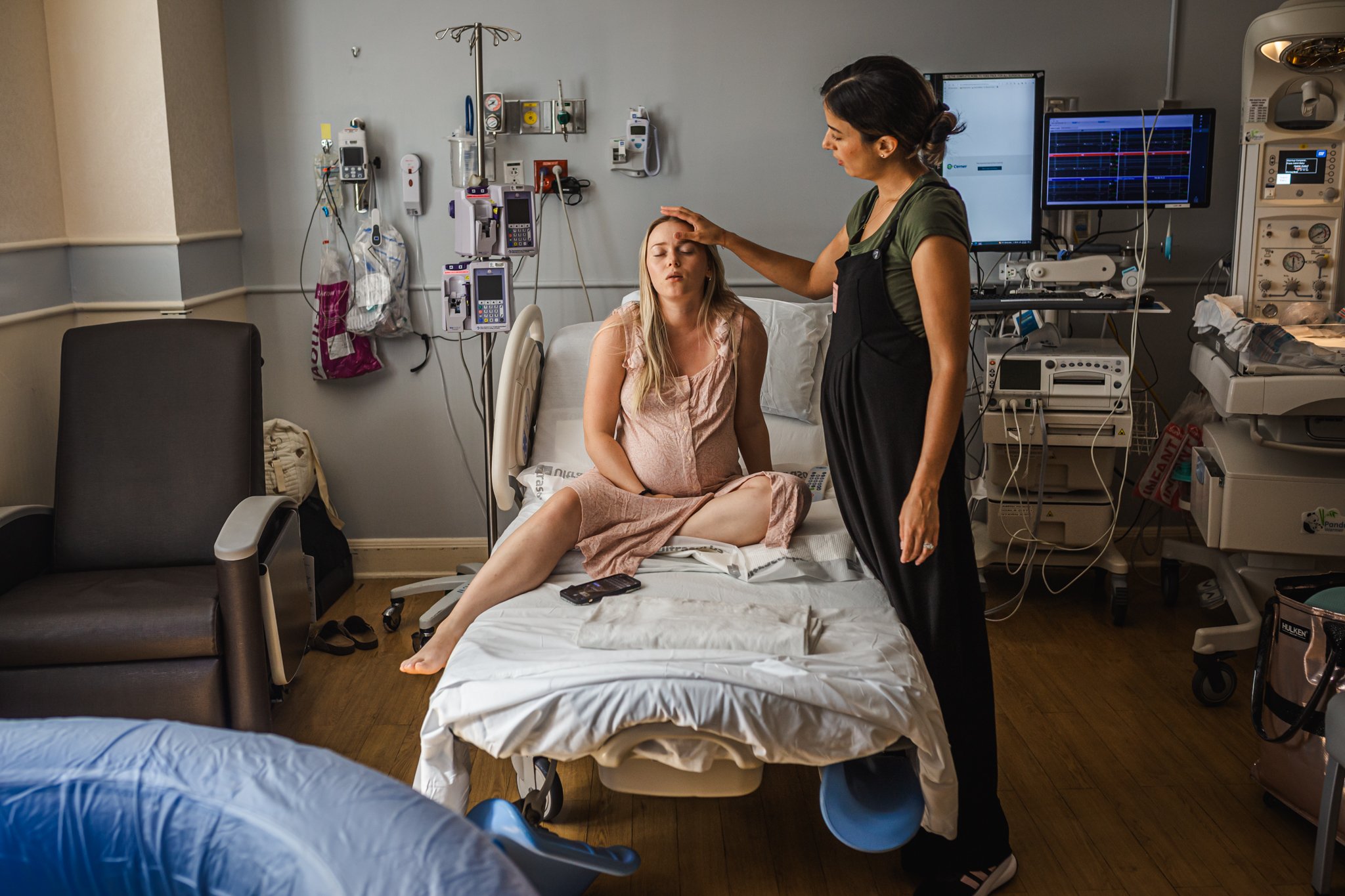 A young woman in a hospital bed with her eyes closed, while a healthcare professional gently touches her forehead, in a hospital room with medical equipment and monitors.