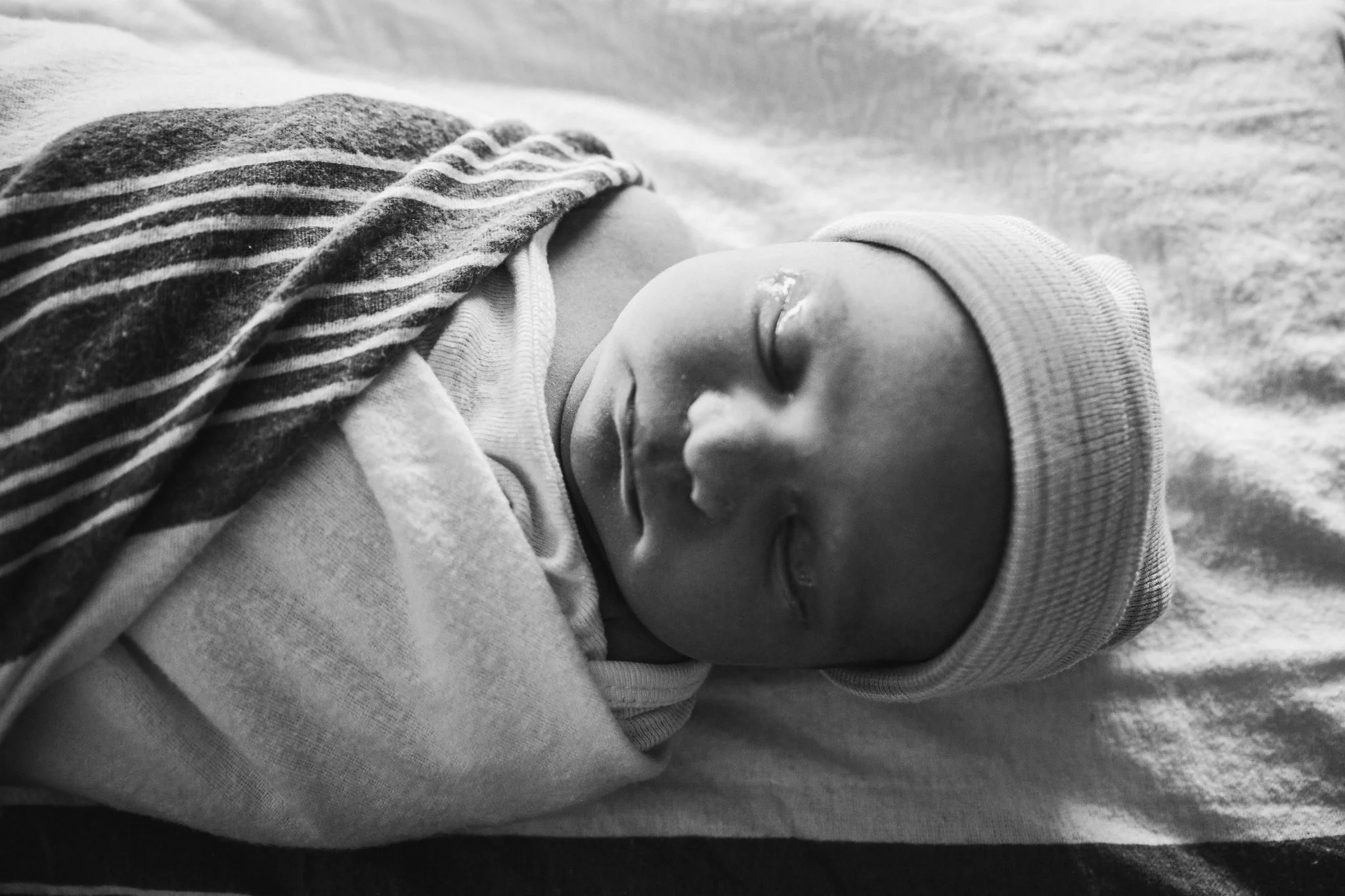 A sleeping baby wearing a striped shirt and a beanie, lying on a soft surface.
