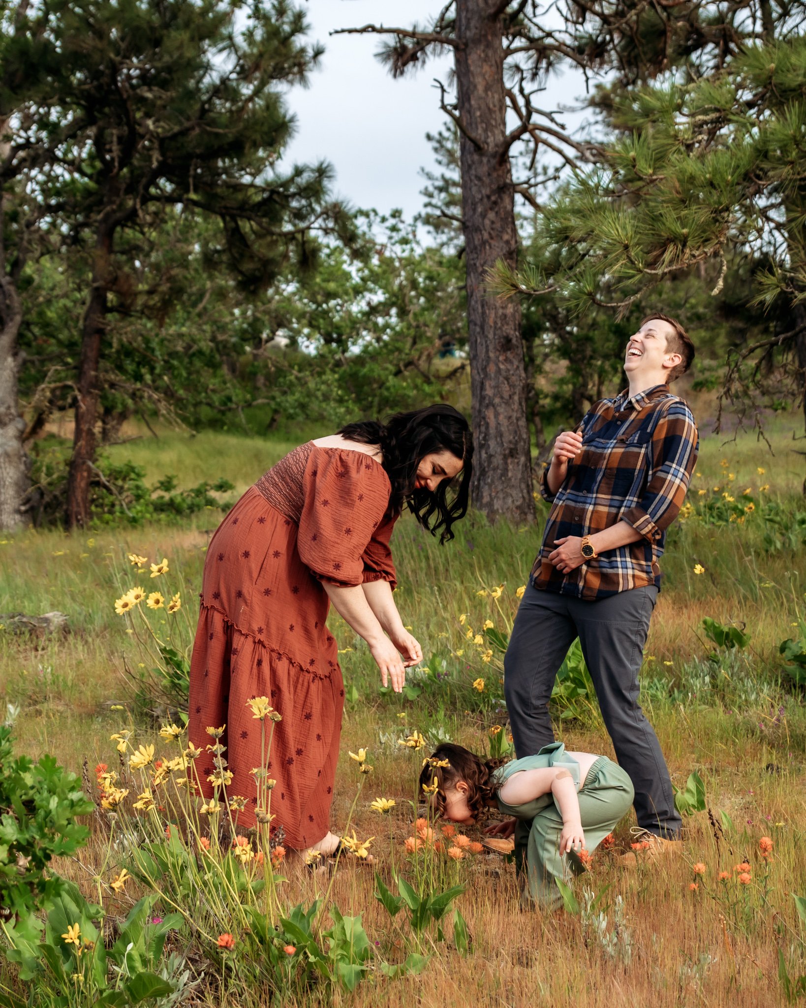 Two women and one girl in a grassy field with yellow and orange flowers, surrounded by pine trees, enjoying a sunny day outdoors.