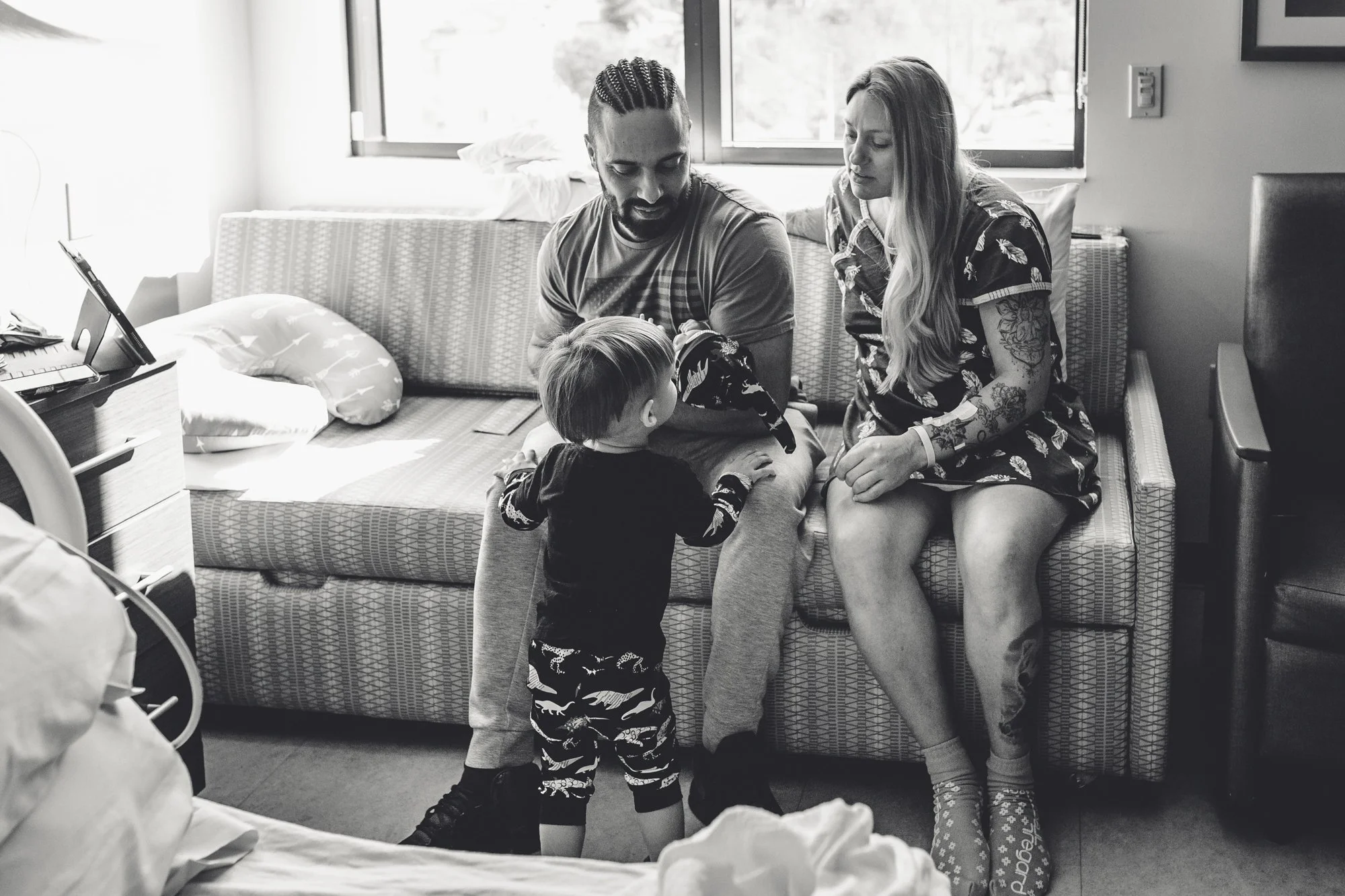 A black and white photo of a family in a hospital room. A father, mother, and young child are sitting on a hospital bed. The father has braided hair and a beard; the mother has long hair with tattoos on her arms. The child is standing and facing the 