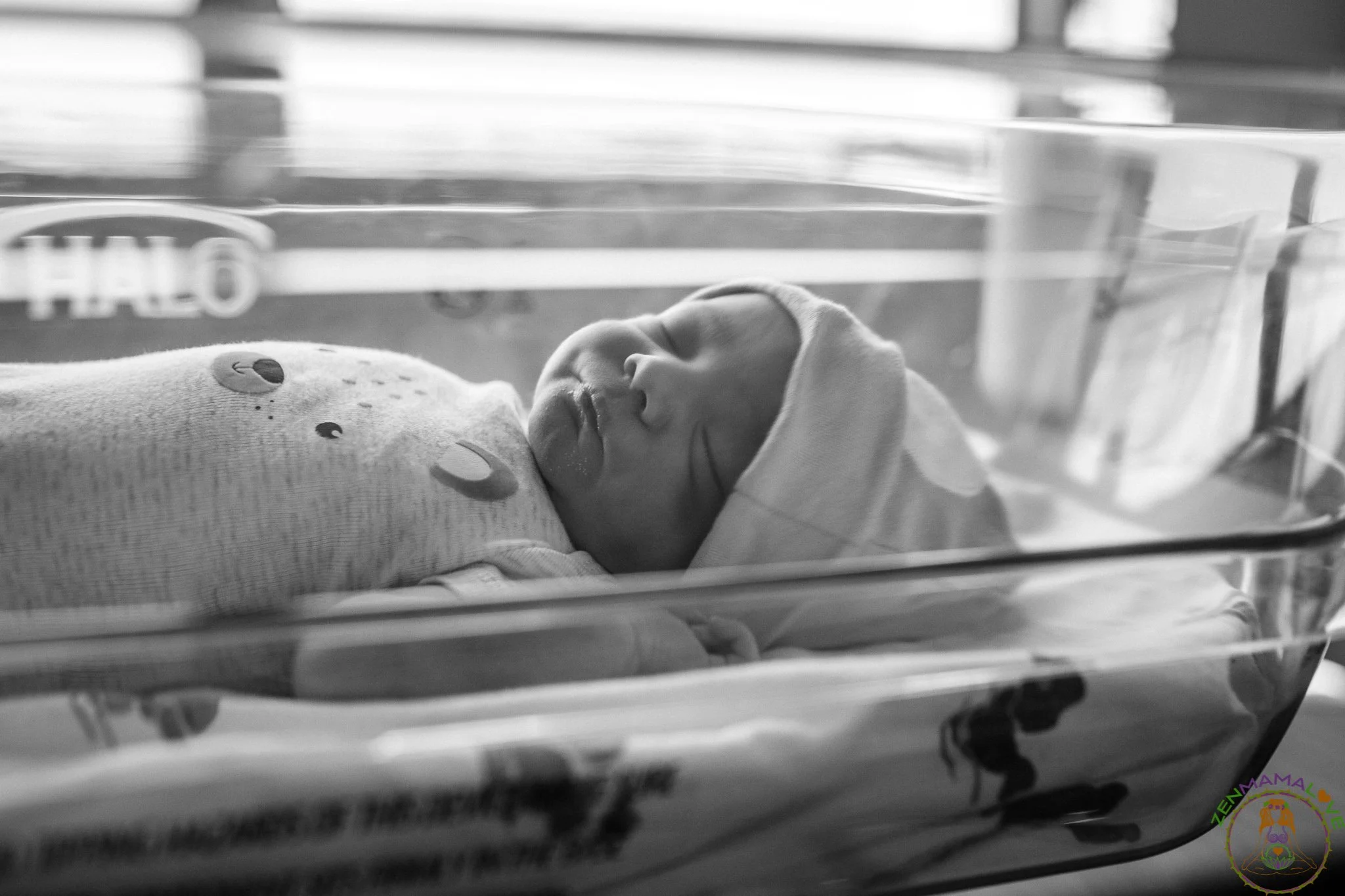 A newborn baby lying in a hospital bassinet with a soft blanket featuring smiley face patterns, resting with a hand near its face in a hospital setting.