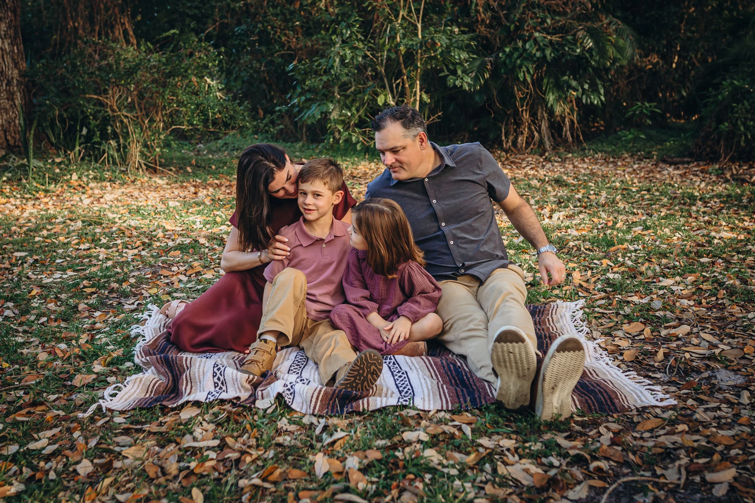 A family of four sitting on a patterned blanket on the ground in a forest, surrounded by fallen leaves and green foliage, enjoying a moment together.