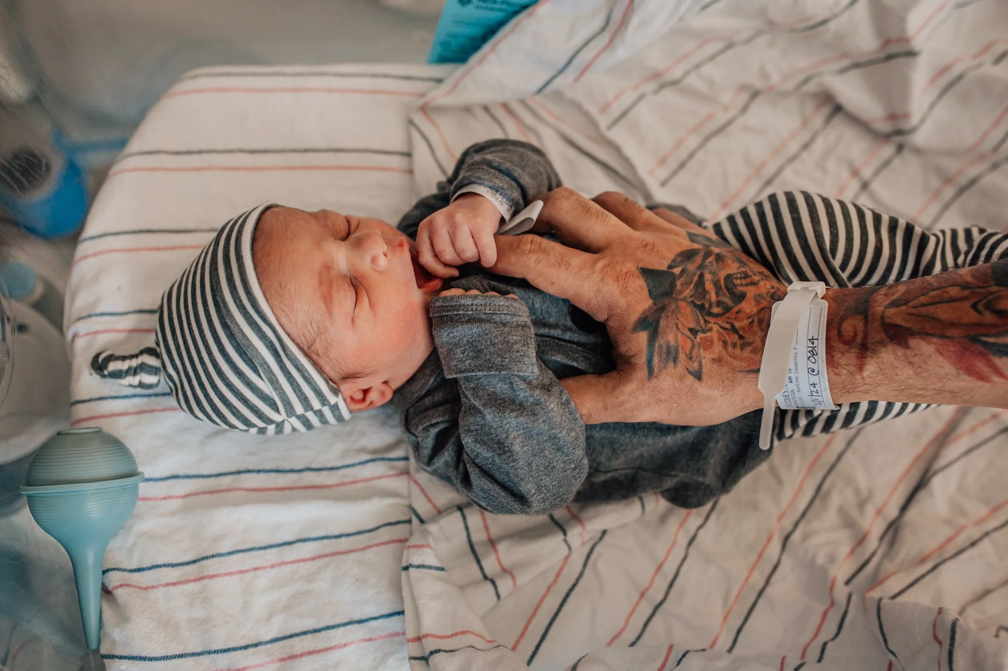 A newborn baby lying in a hospital crib, wearing a striped hat and pajamas, holding an adult's finger. The adult has tattoos on their arm and is wearing a hospital wristband. There's a blue suction device nearby.