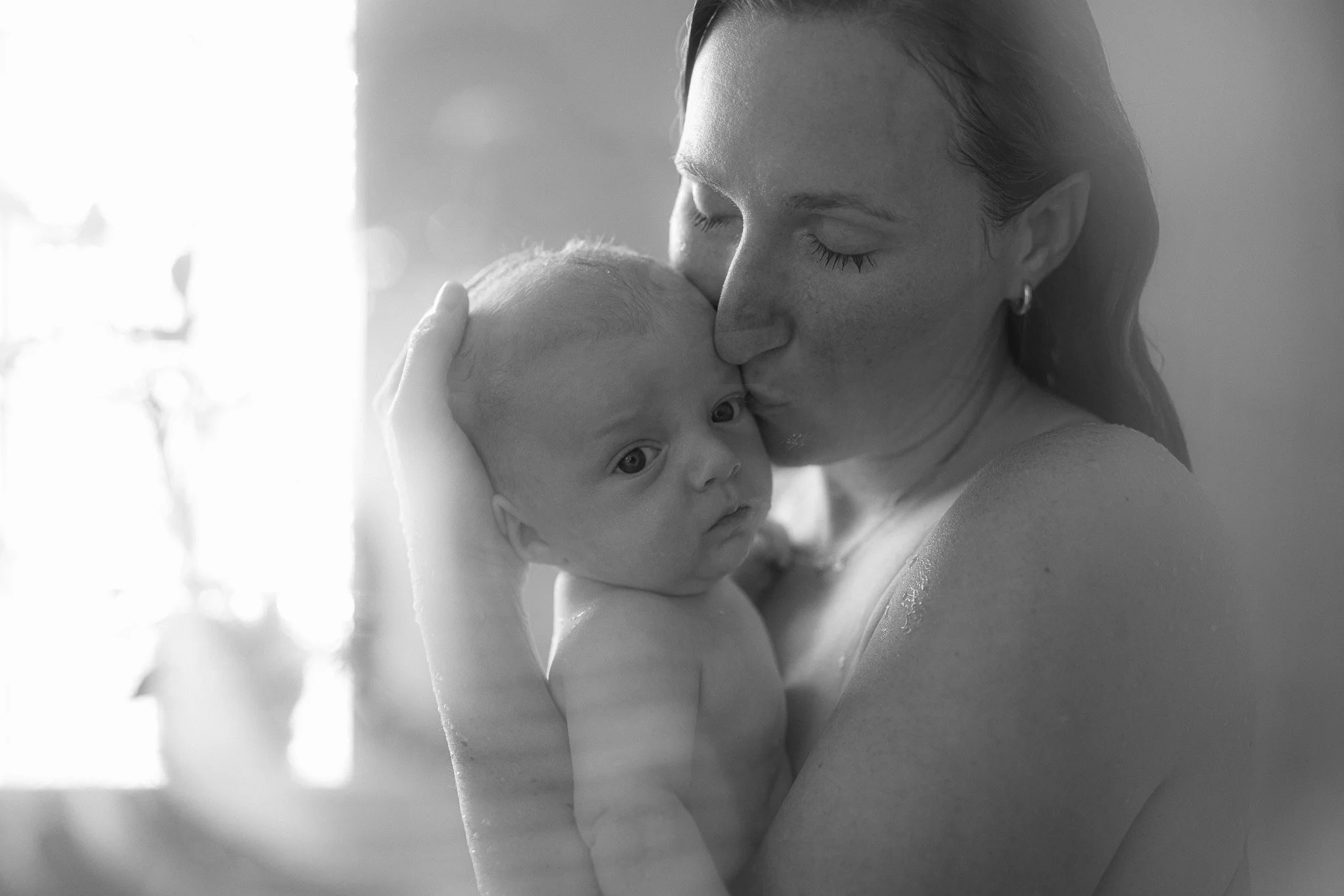 A woman holding and kissing a newborn baby in black and white, with soft-lit background.