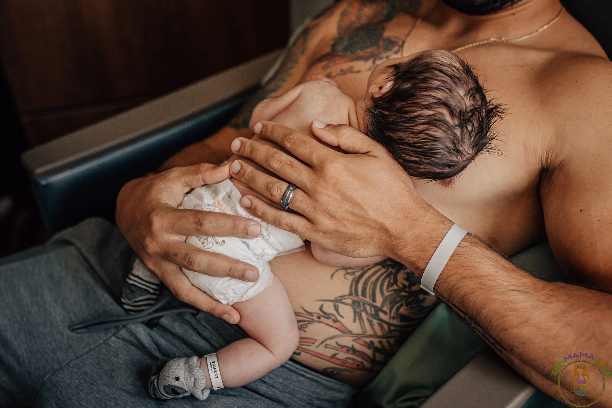 A newborn baby breastfeeding from a tattooed man, both with hospital wristbands, the man lying on a hospital bed.