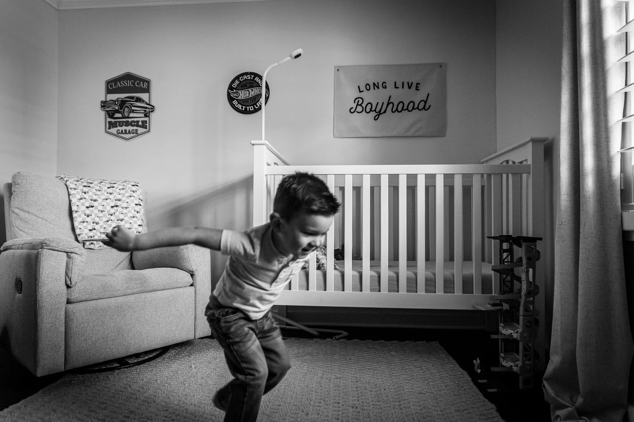 A young boy with dark hair playing with a toy sword in a living room with a sofa, patterned blanket, and a nursery or crib in the background, along with wall decorations and a window.